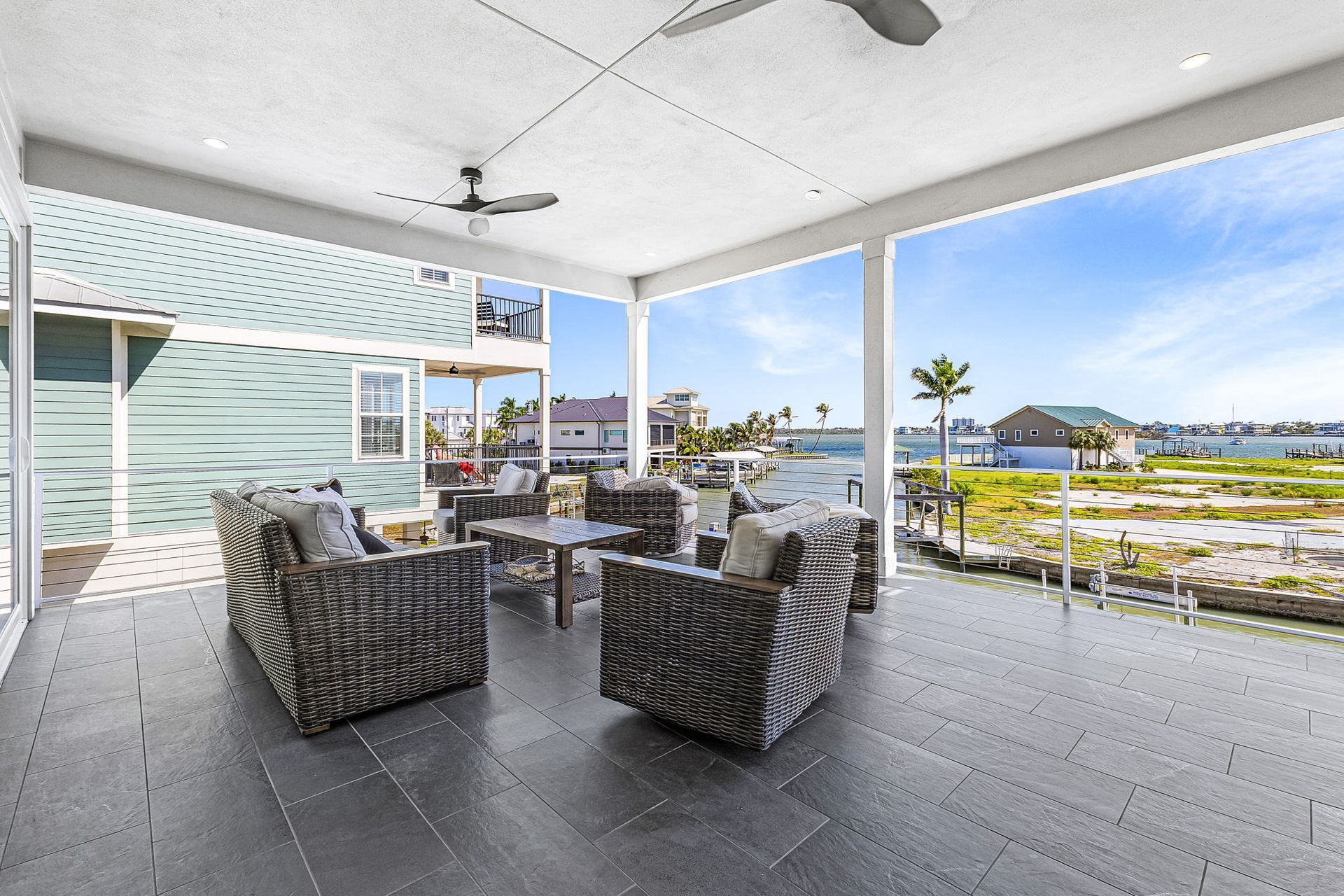 Covered waterfront patio with wicker seating, tables, and ocean views under a white roof.