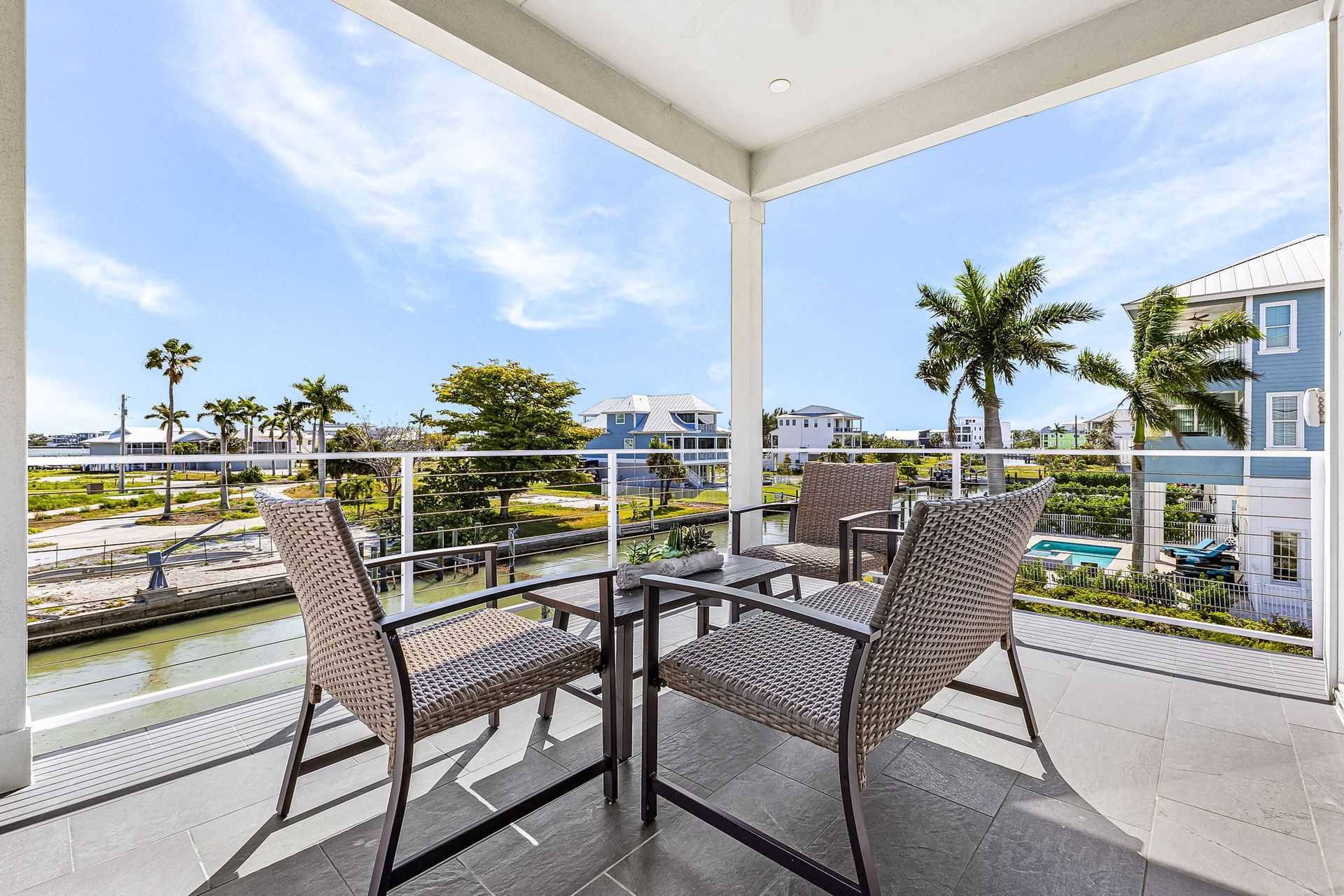 Covered balcony with wicker chairs and table overlooking palm trees, water, and blue seaside buildings.