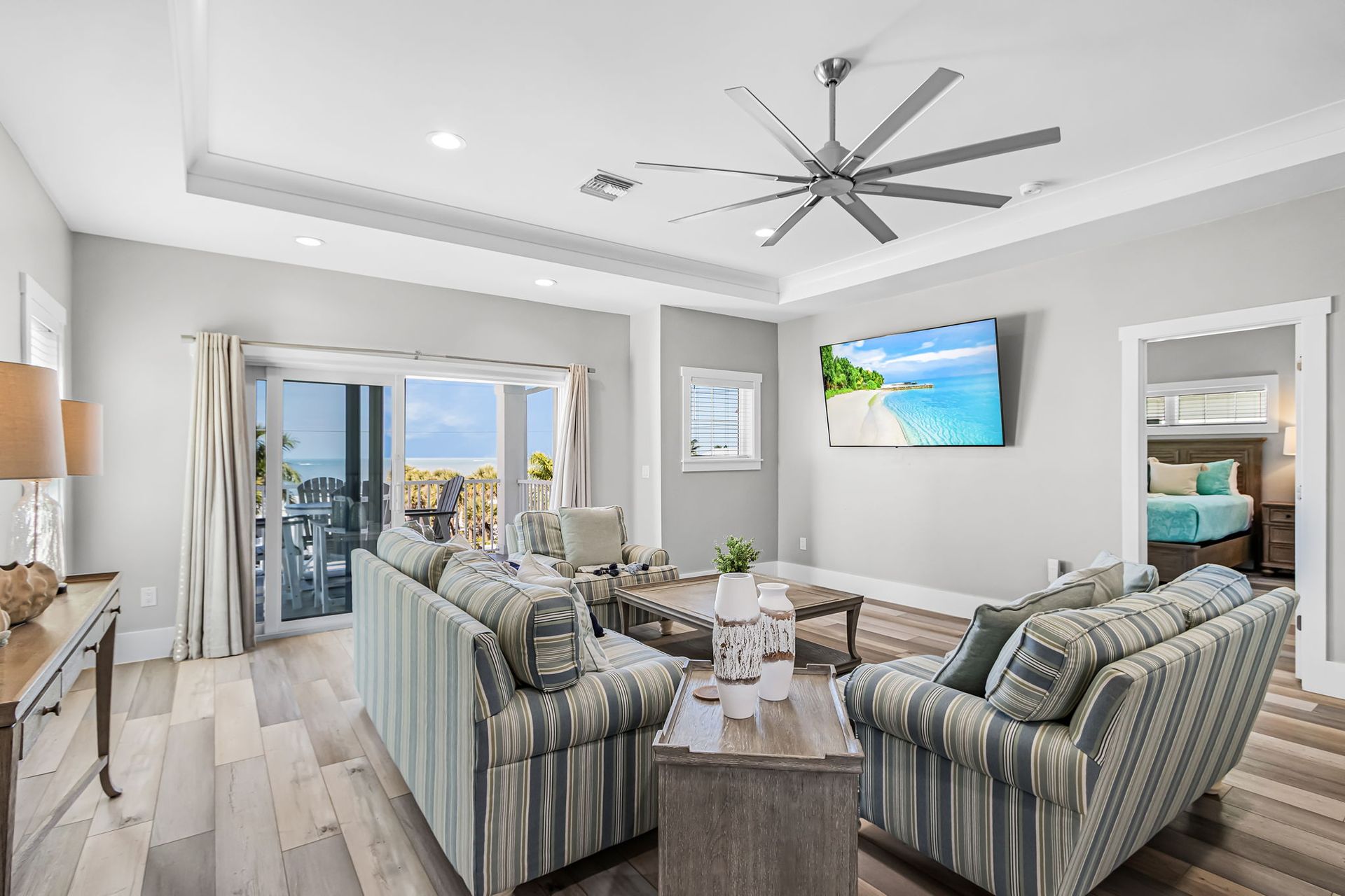 Bright living room with striped sofas, wall-mounted TV, ceiling fan, and balcony view through sliding glass doors