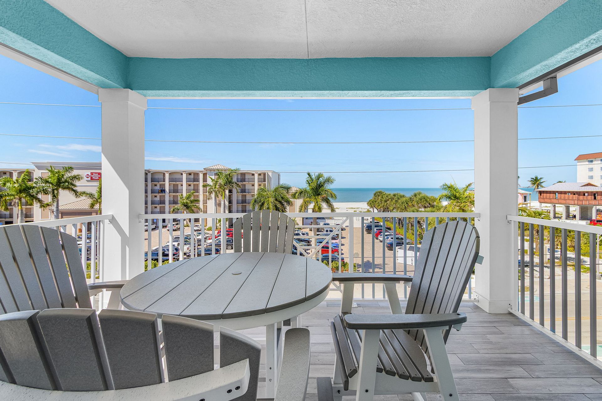Covered balcony with white patio table and chairs overlooking palm trees, buildings, and the ocean.