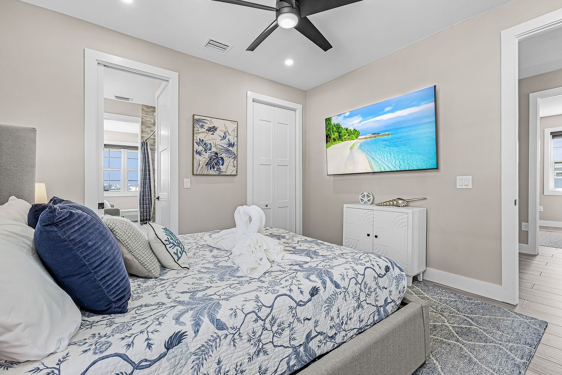 Bright bedroom with a bed, wall-mounted TV, ceiling fan, and beach artwork.