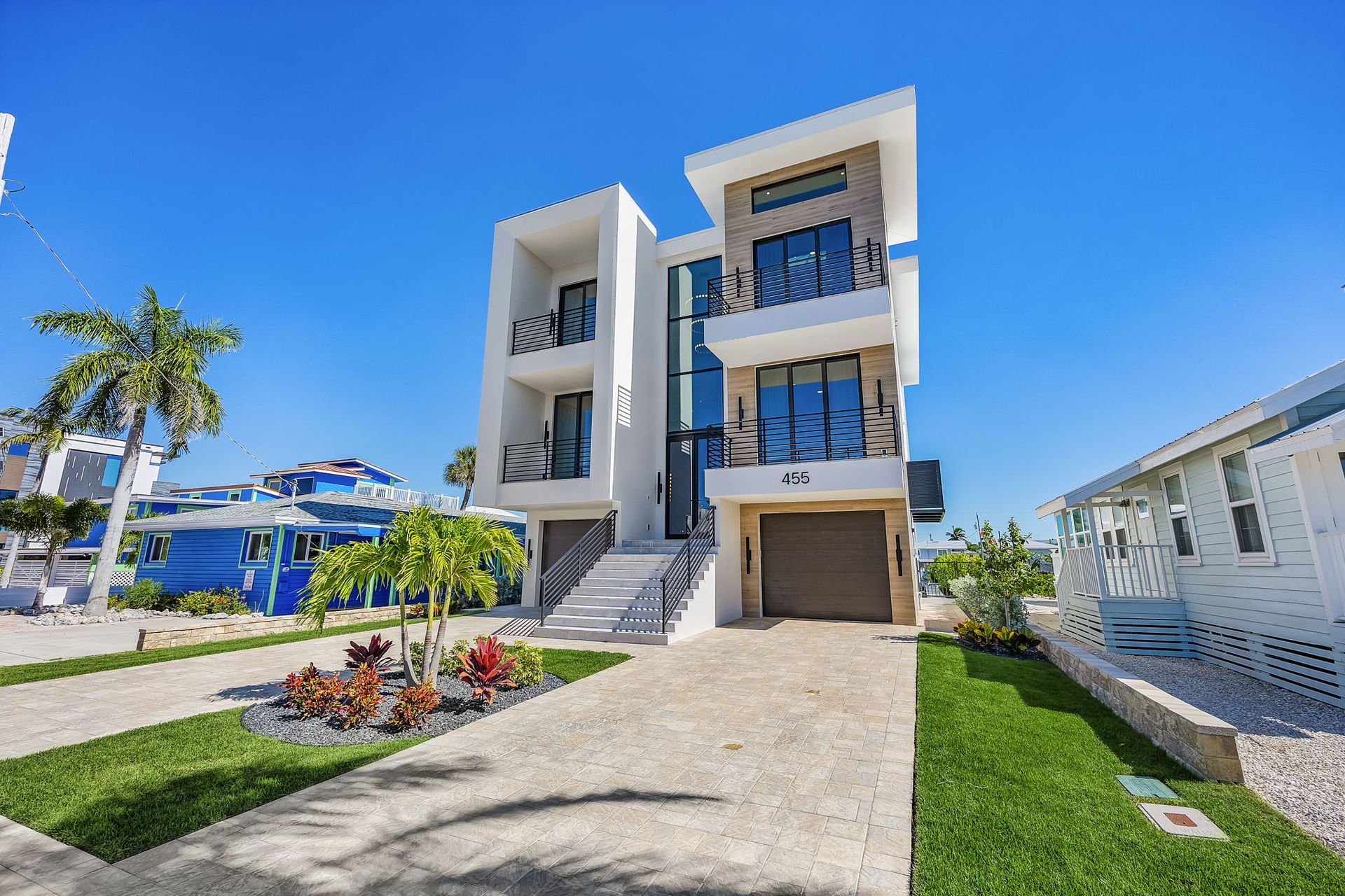 Modern white multi-story house with garage, steps, and palm trees under a blue sky.