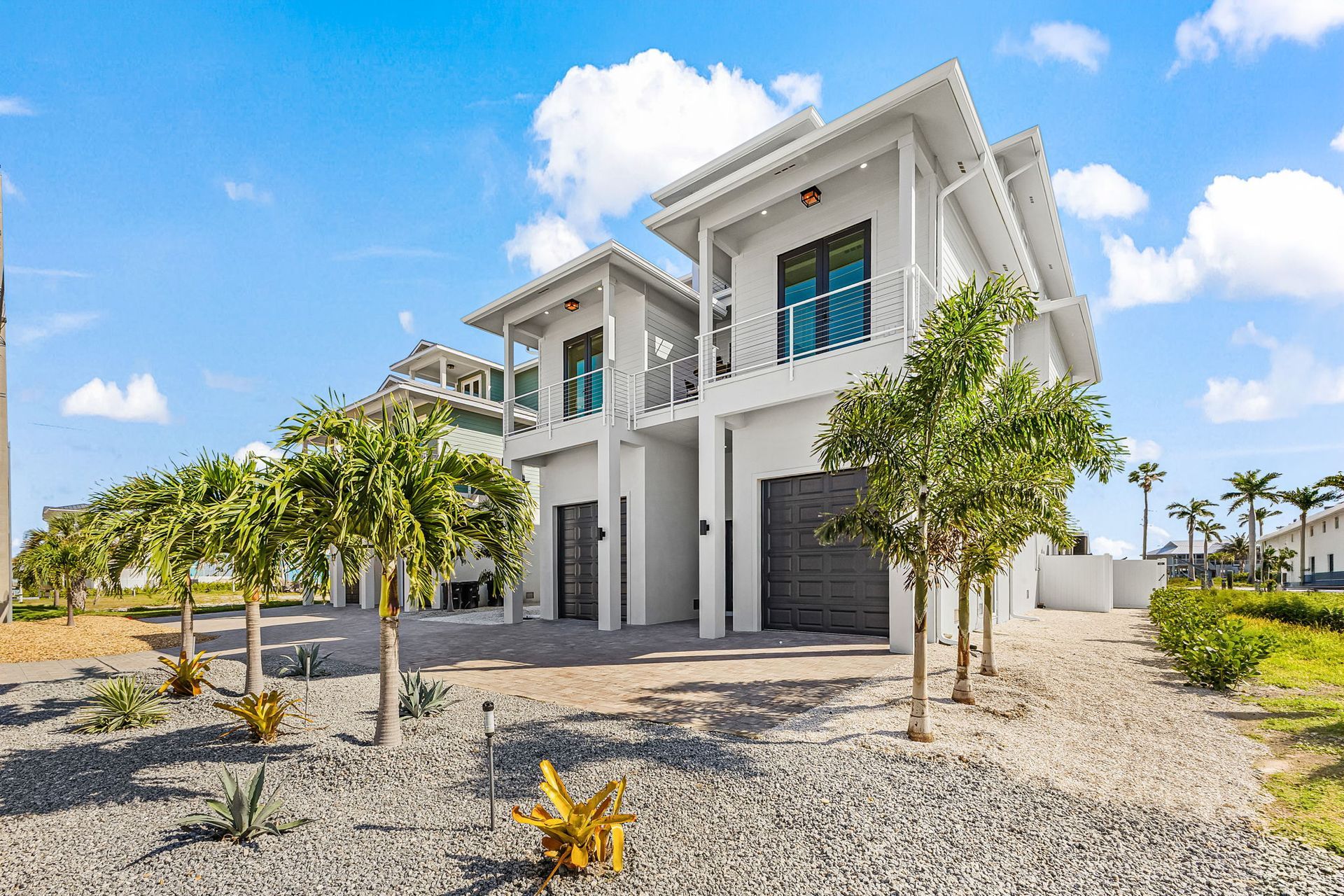 Modern white two-story house with large windows, palm trees, and a gravel front yard under a blue sky