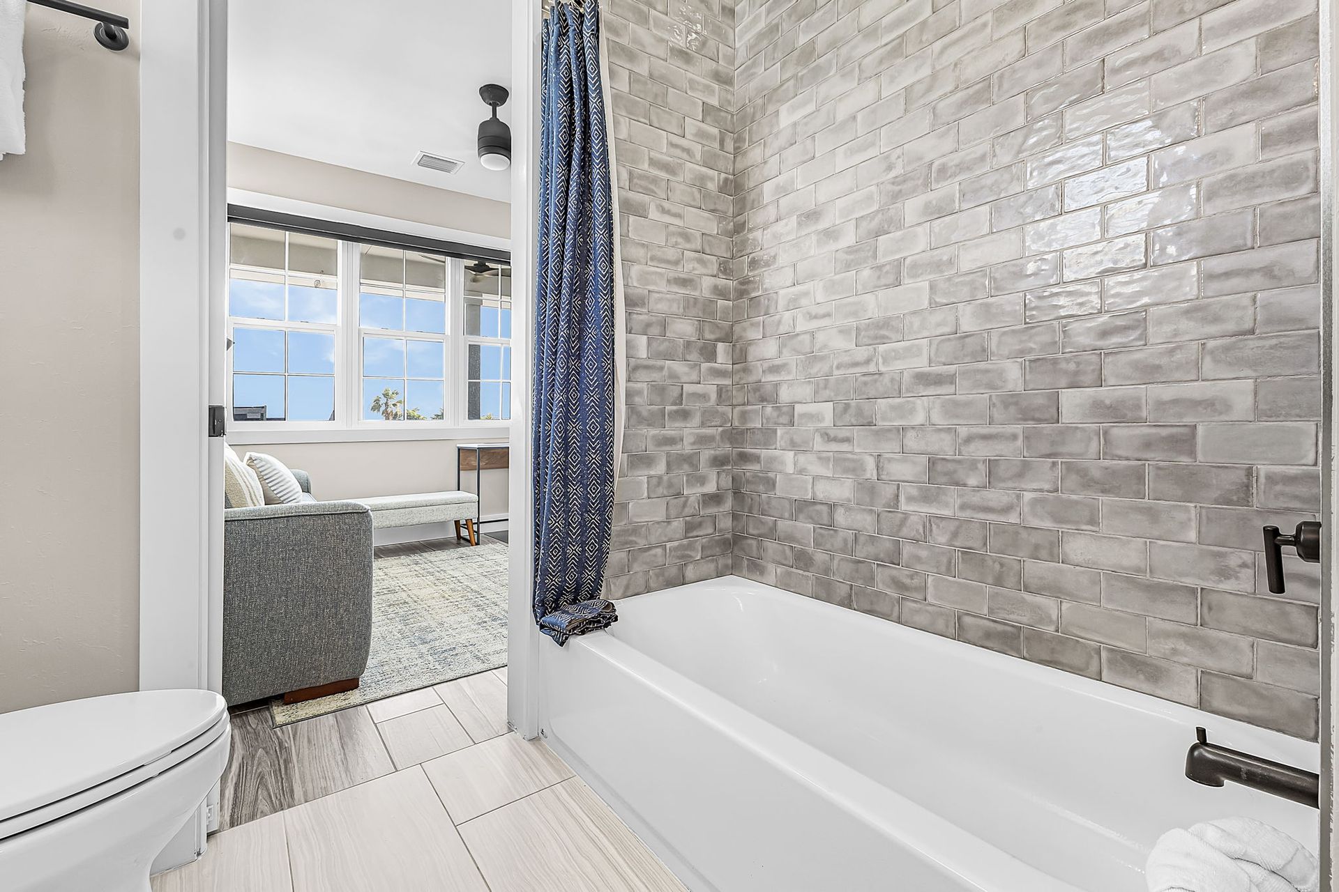 Bright bathroom with tiled walls, white tub, and curtain, opening to a bedroom with a window.