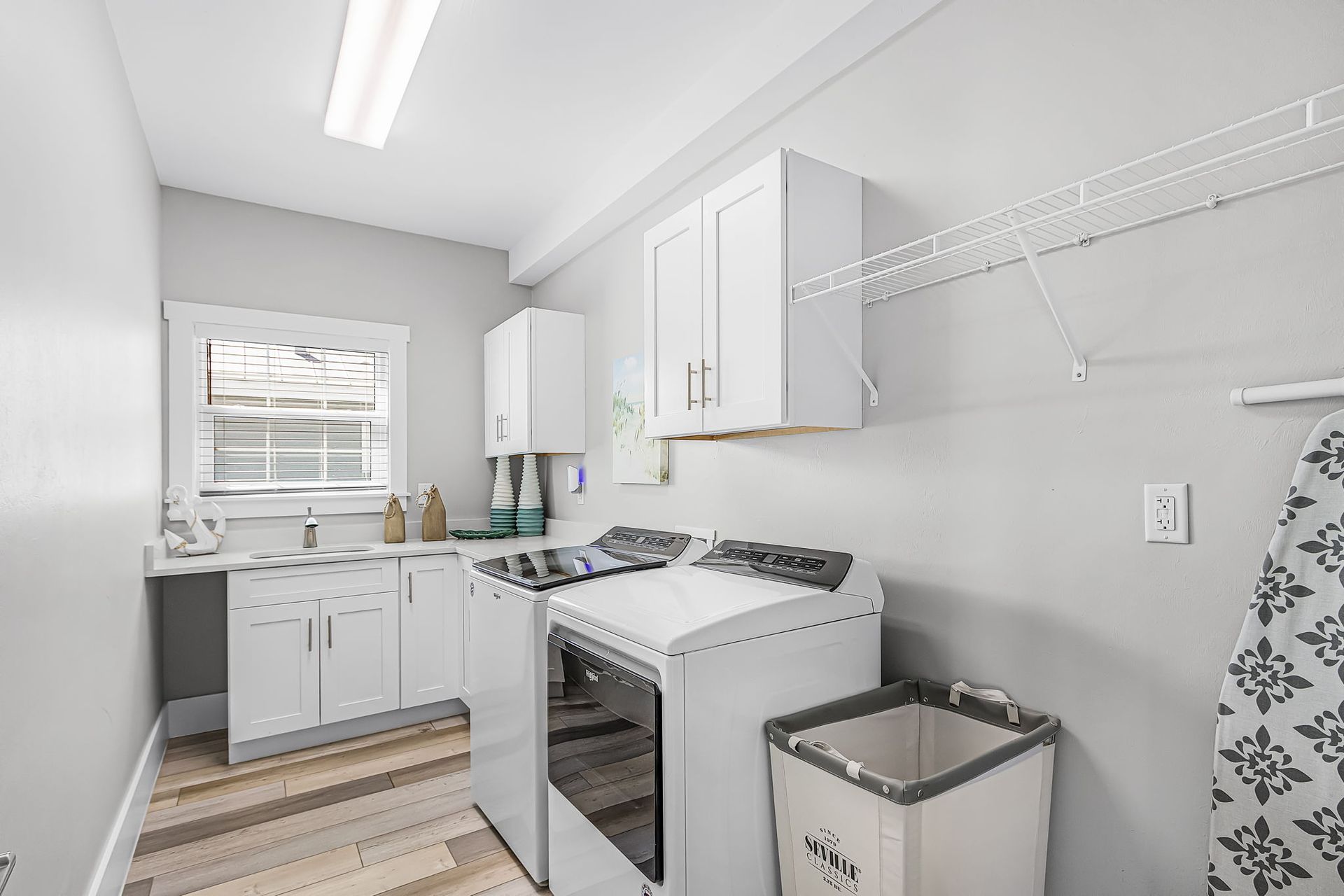 Bright laundry room with white cabinets, washer, dryer, sink, and wire shelving.