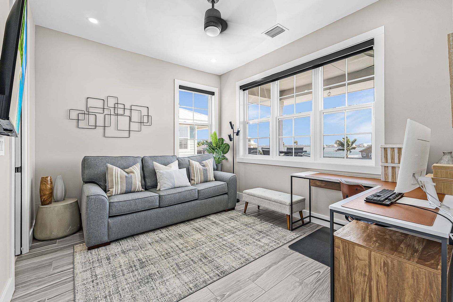 Bright home office with a gray sofa, desk, large windows, and a light patterned rug.