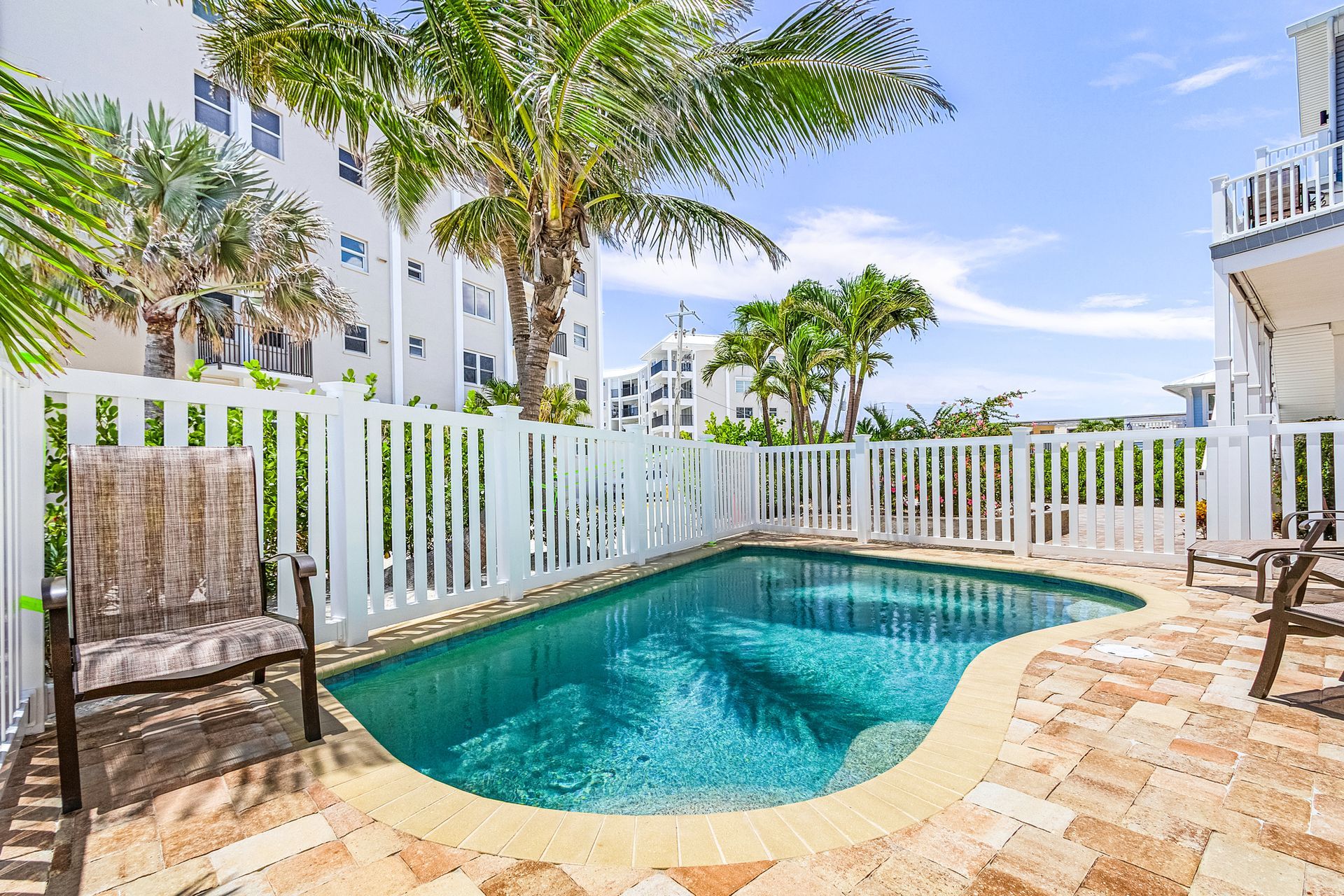 Small turquoise backyard pool beside a white fence, palm trees, and a wicker chair on a sunlit patio