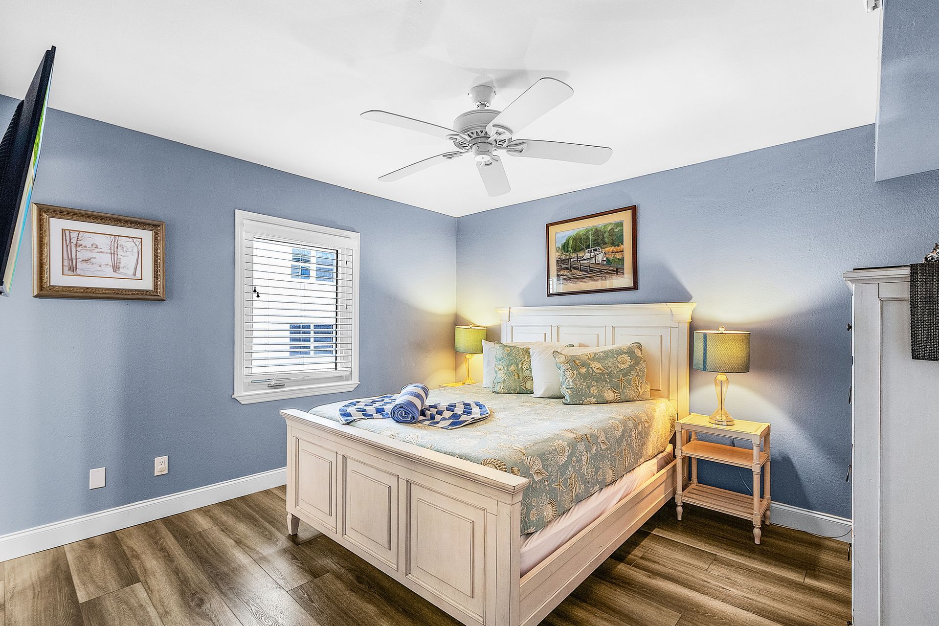 Blue bedroom with a white bed, bedside lamp, framed art, and a ceiling fan