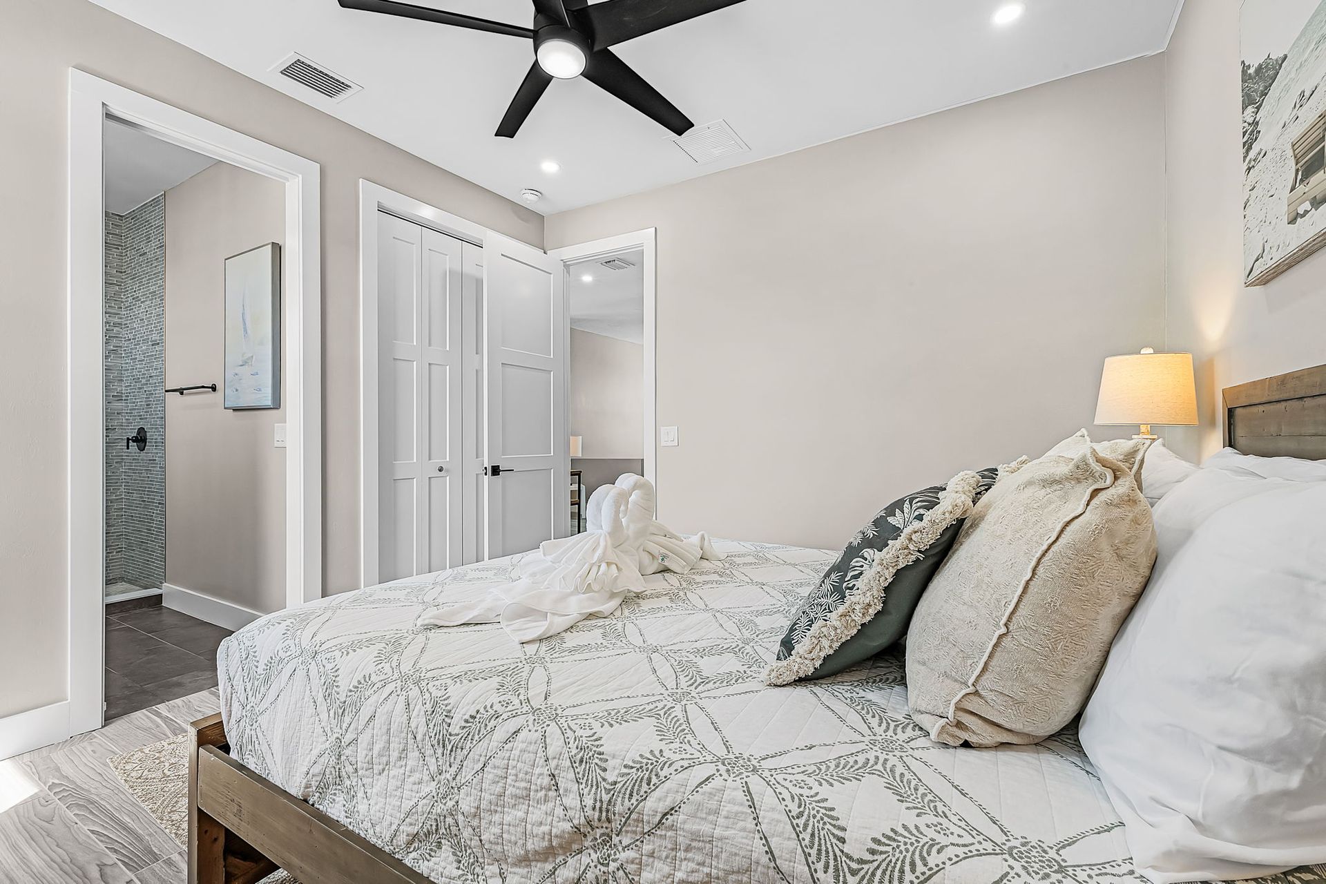 Bright bedroom with a made bed, ceiling fan, mirrored closet doors, and a lit bedside lamp