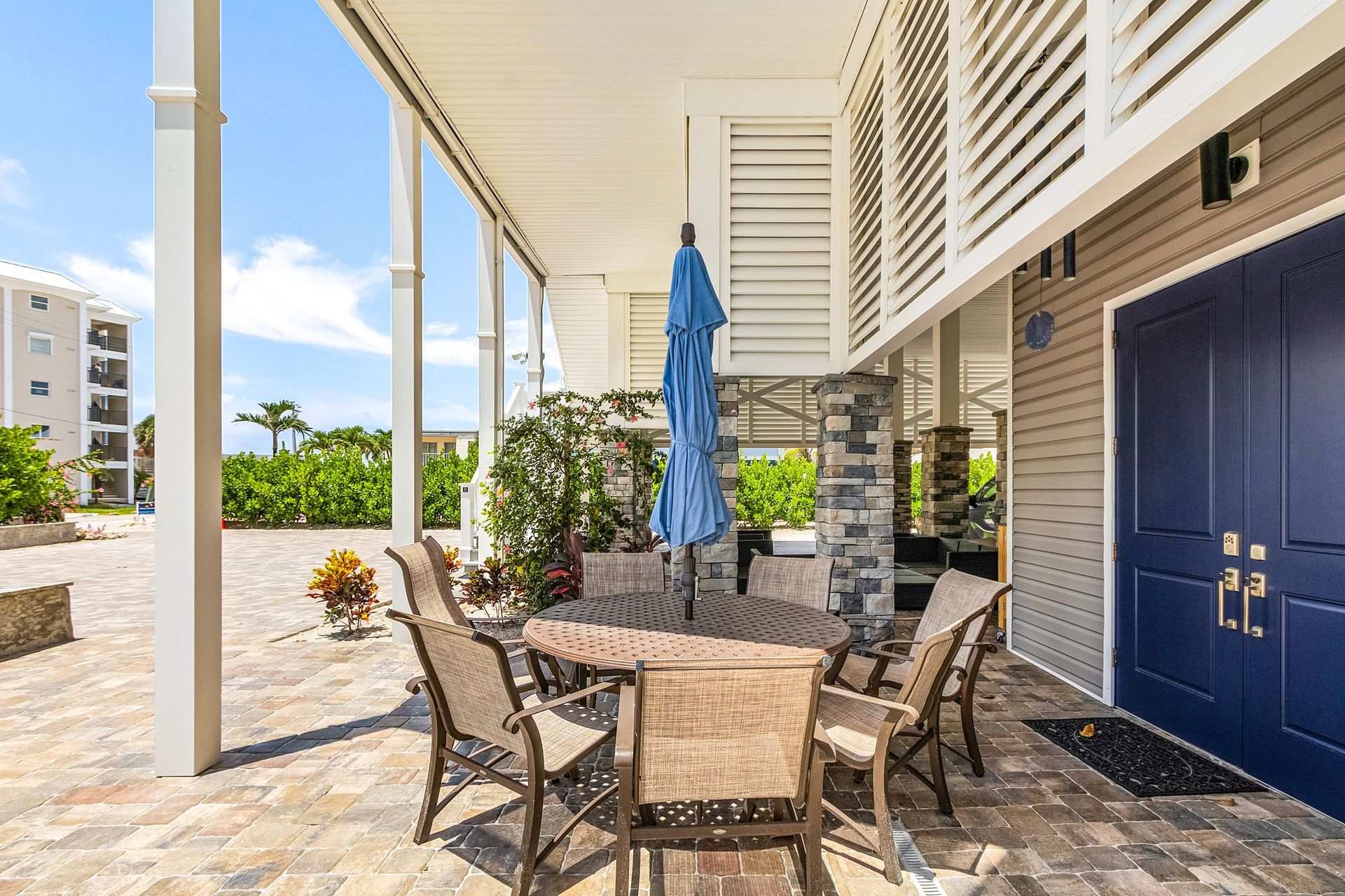Patio dining area with wicker chairs, round table, blue umbrella, and navy double doors beside a poolside walkway