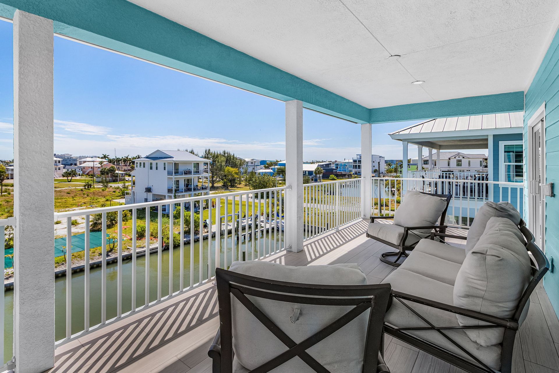 Covered balcony with white railings, turquoise trim, and patio chairs overlooking the water and nearby homes