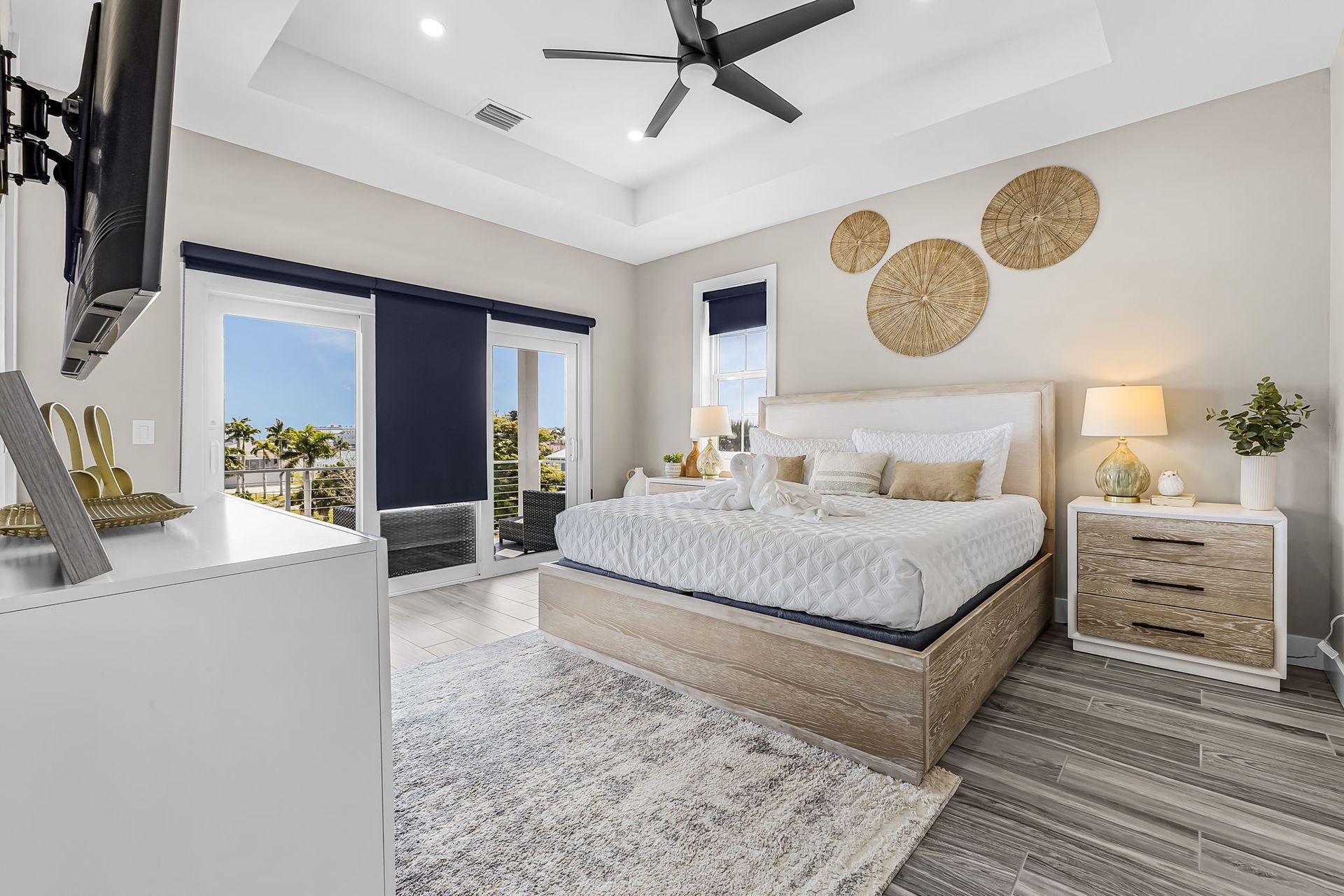 Bright modern bedroom with king bed, wicker nightstand, rug, ceiling fan, and balcony doors overlooking palm trees