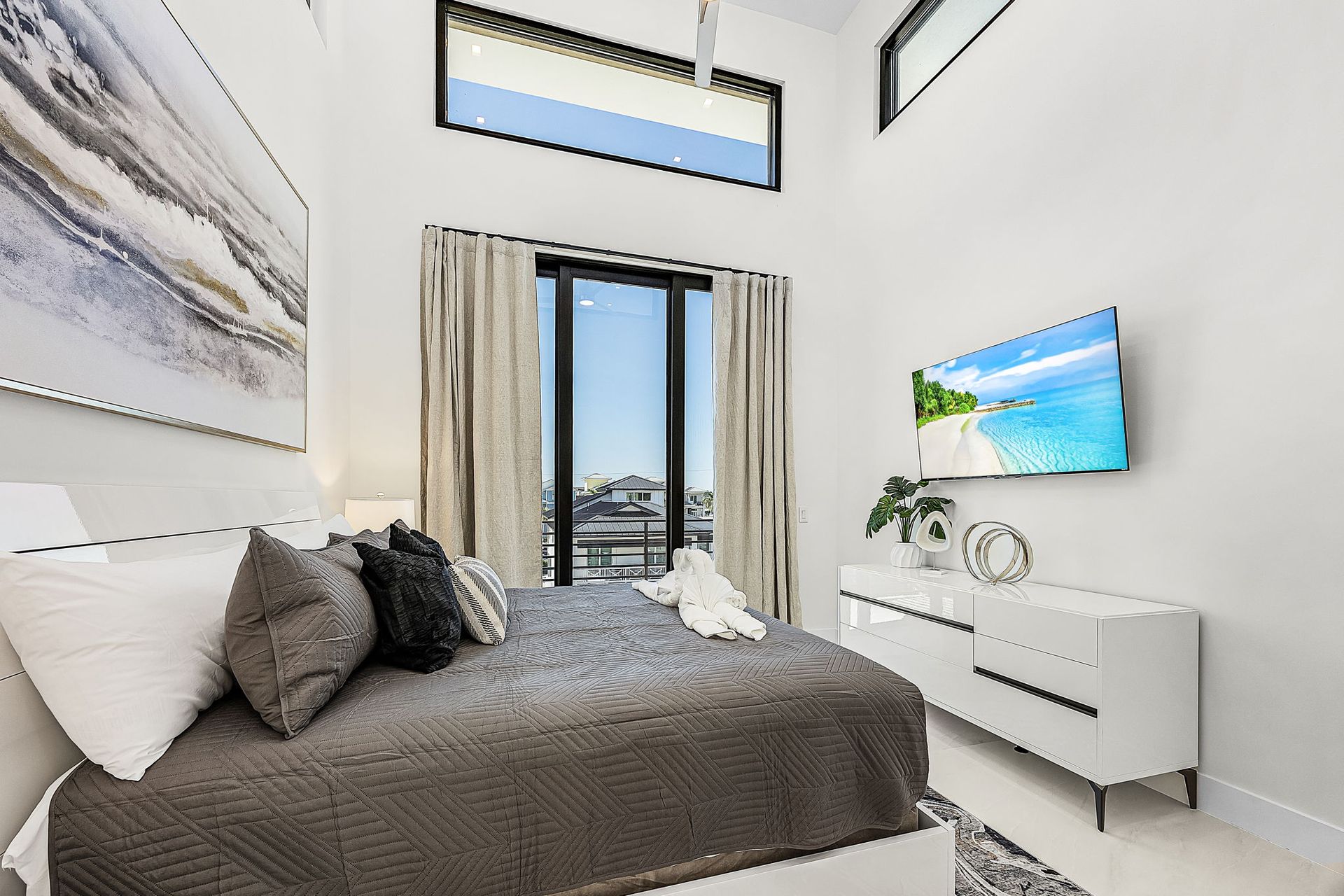 Modern bedroom with a bed, TV, and large window; white walls, gray bedding, and beach scene on TV.