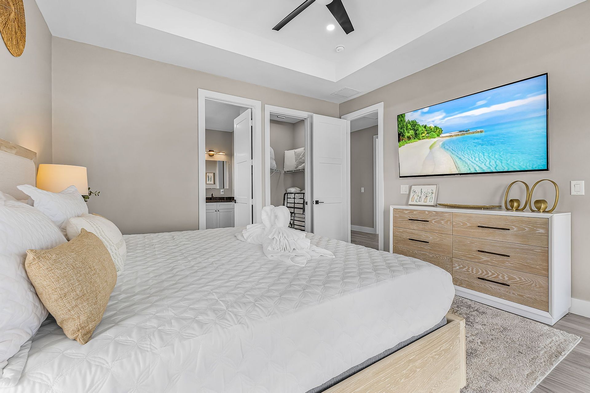 Bright bedroom with a white bed, beige dresser, wall TV, and open doors to a bathroom/closet.