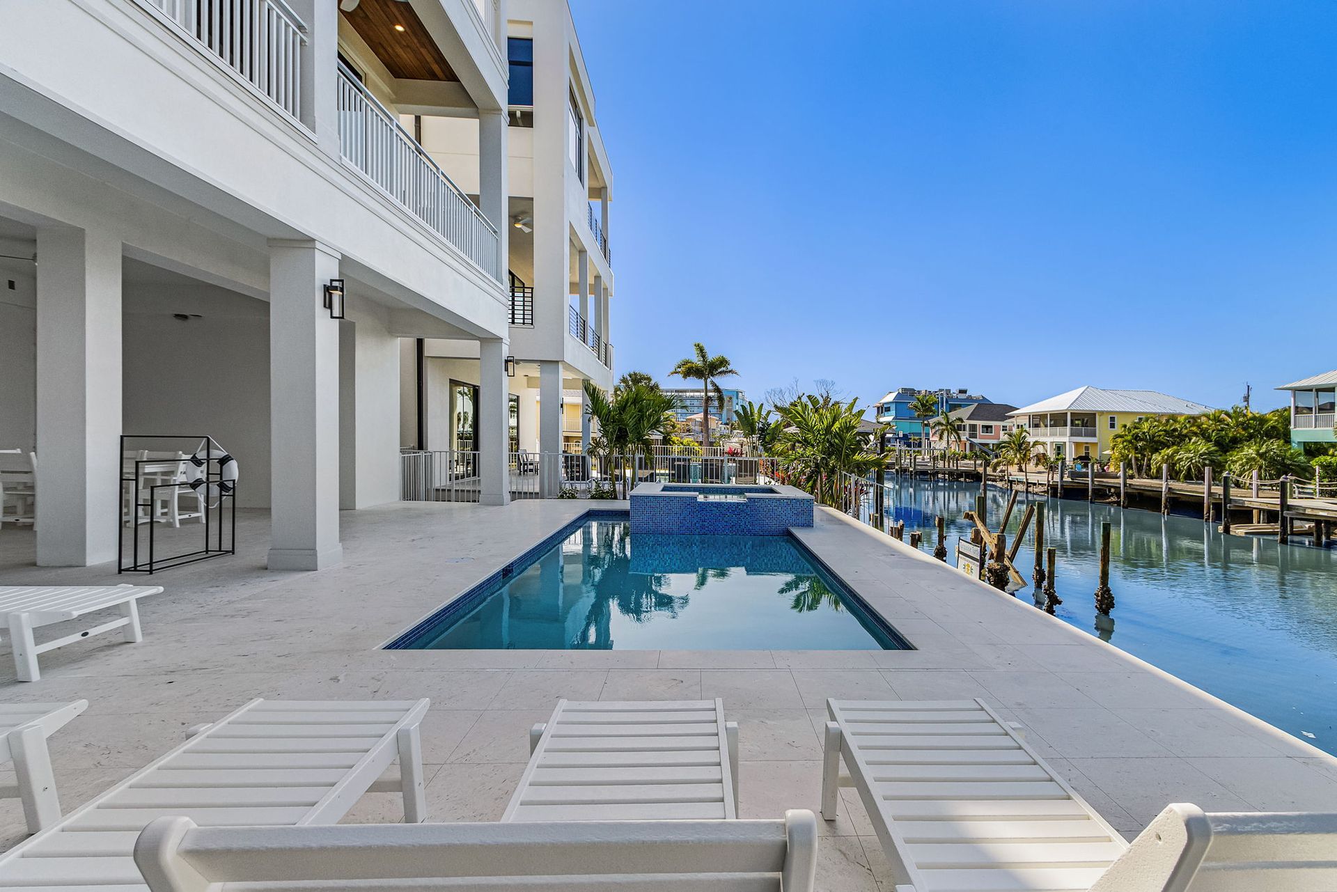 White luxury home with pool overlooking a canal. Blue water, palm trees, and lounge chairs. Sunny day.