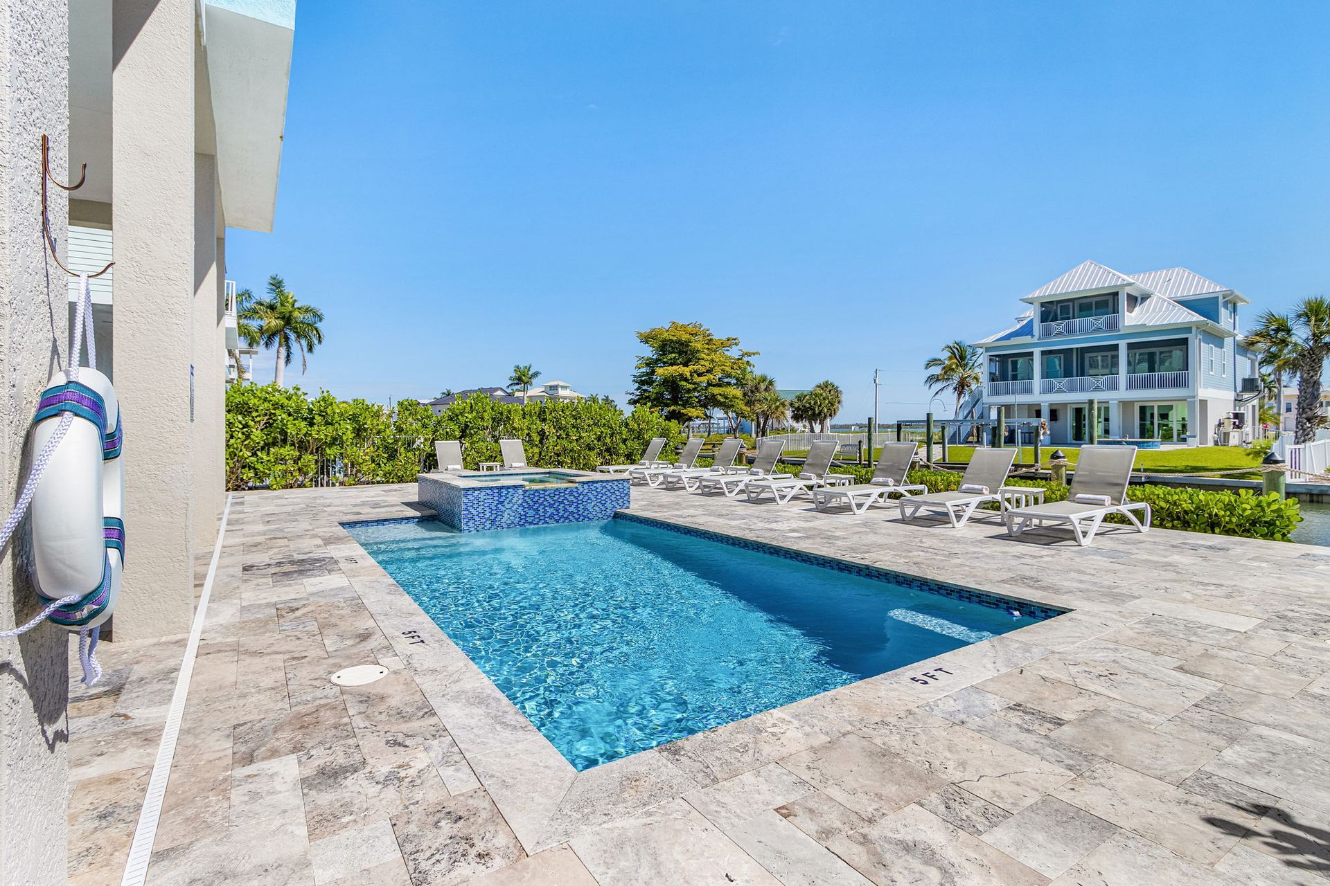 Outdoor pool with blue water, stone patio, lounge chairs, and a waterfront house in the background