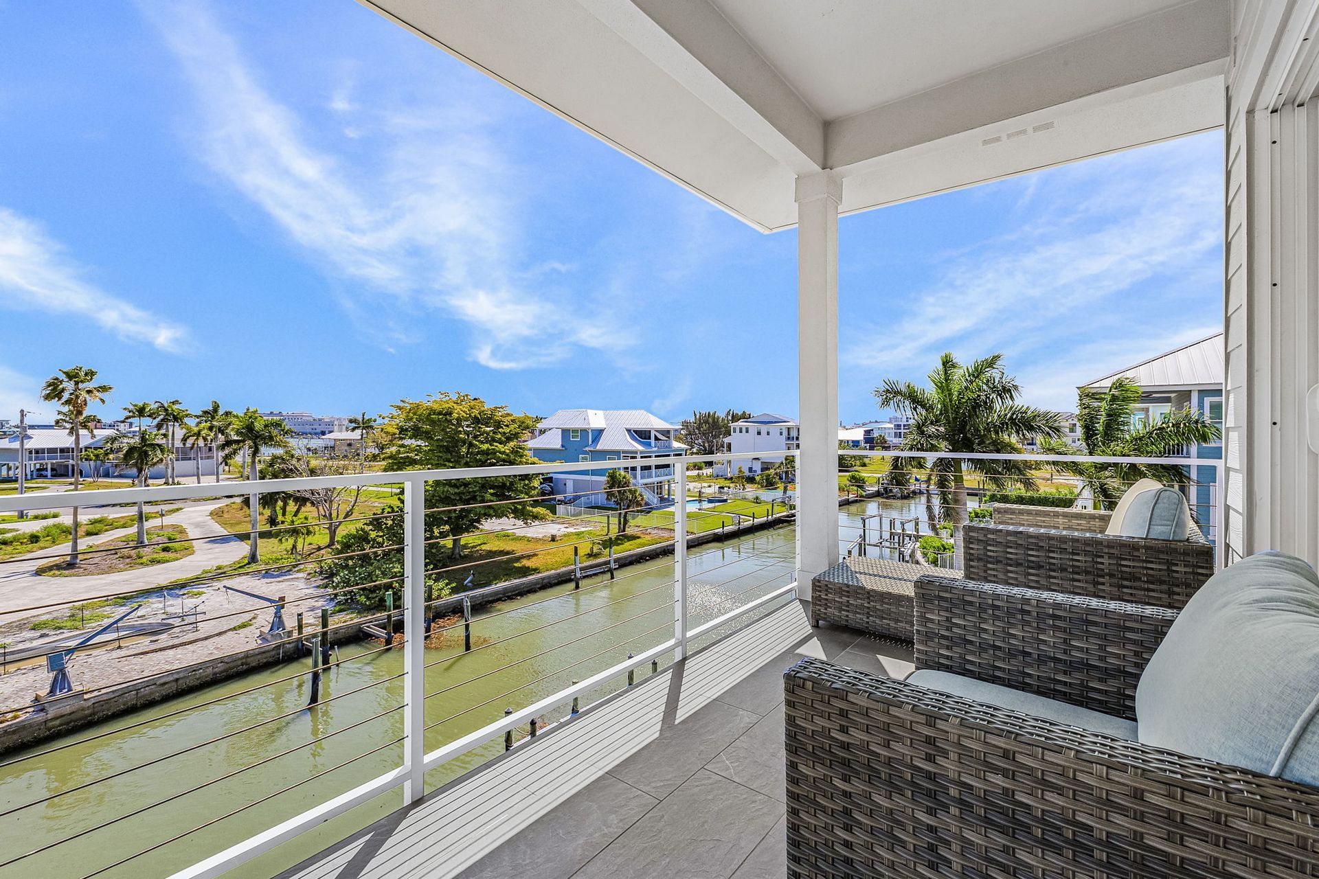 Balcony view of a canal and palm-lined neighborhood, with wicker chairs and glass railing under a blue sky