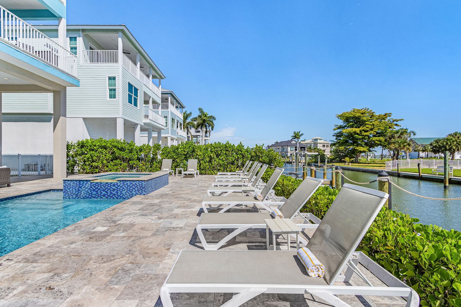 Waterfront resort patio with white lounge chairs beside a pool and canal under a blue sky