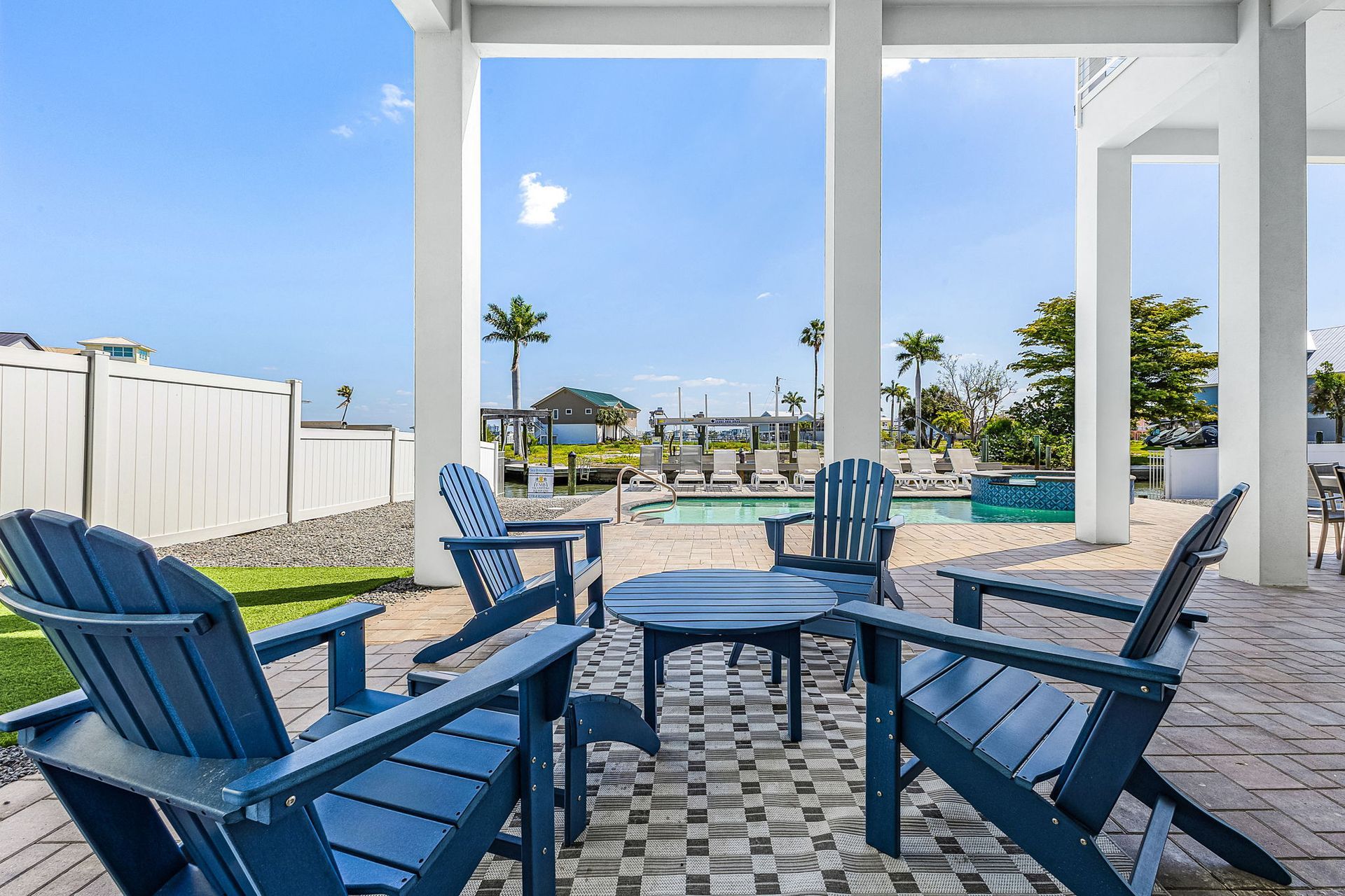 Blue patio chairs and table under white pergola, overlooking a sunny pool area and sky.