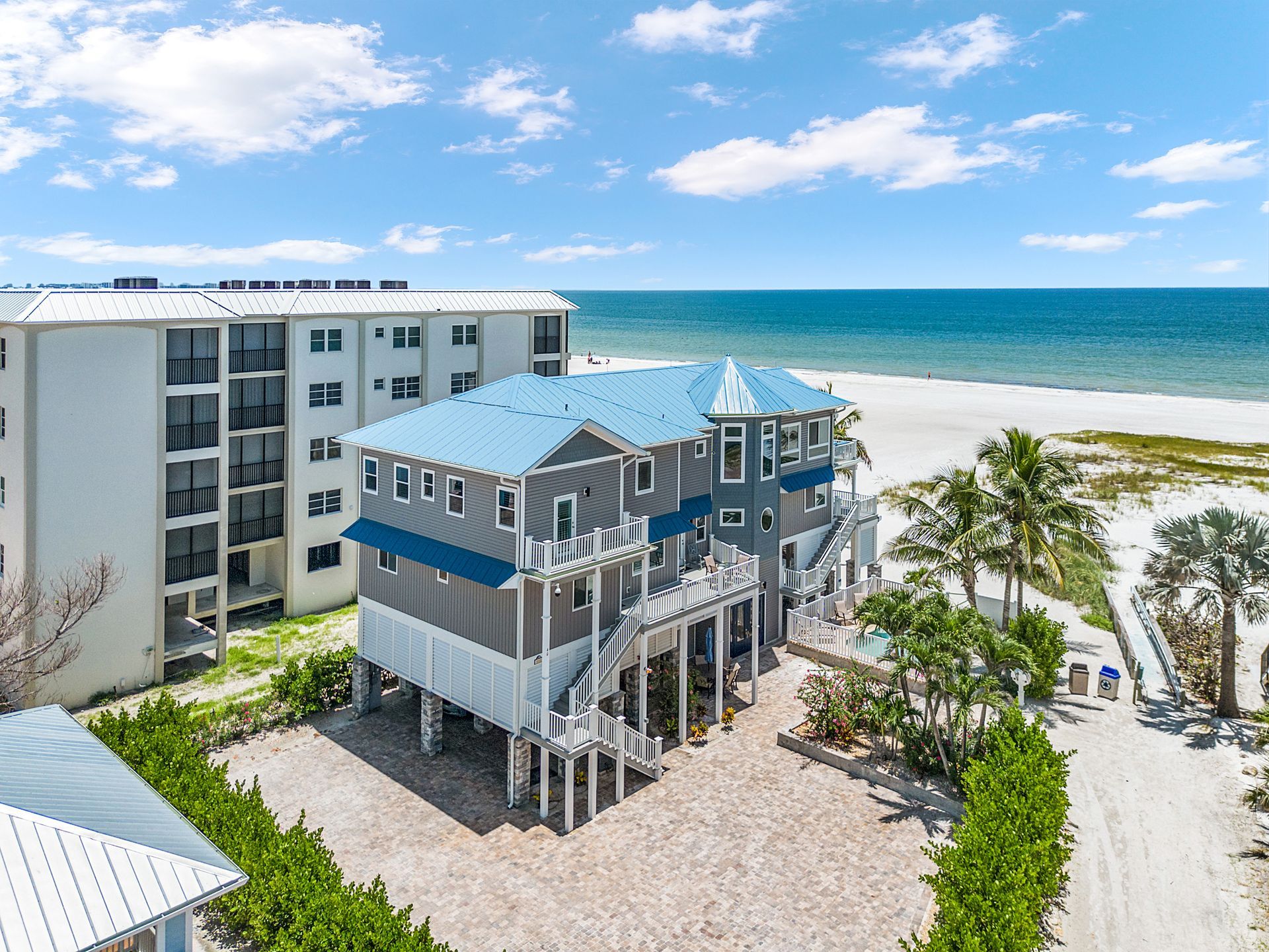 Beachfront condo on stilts beside sand, palm trees, and turquoise ocean under a blue sky