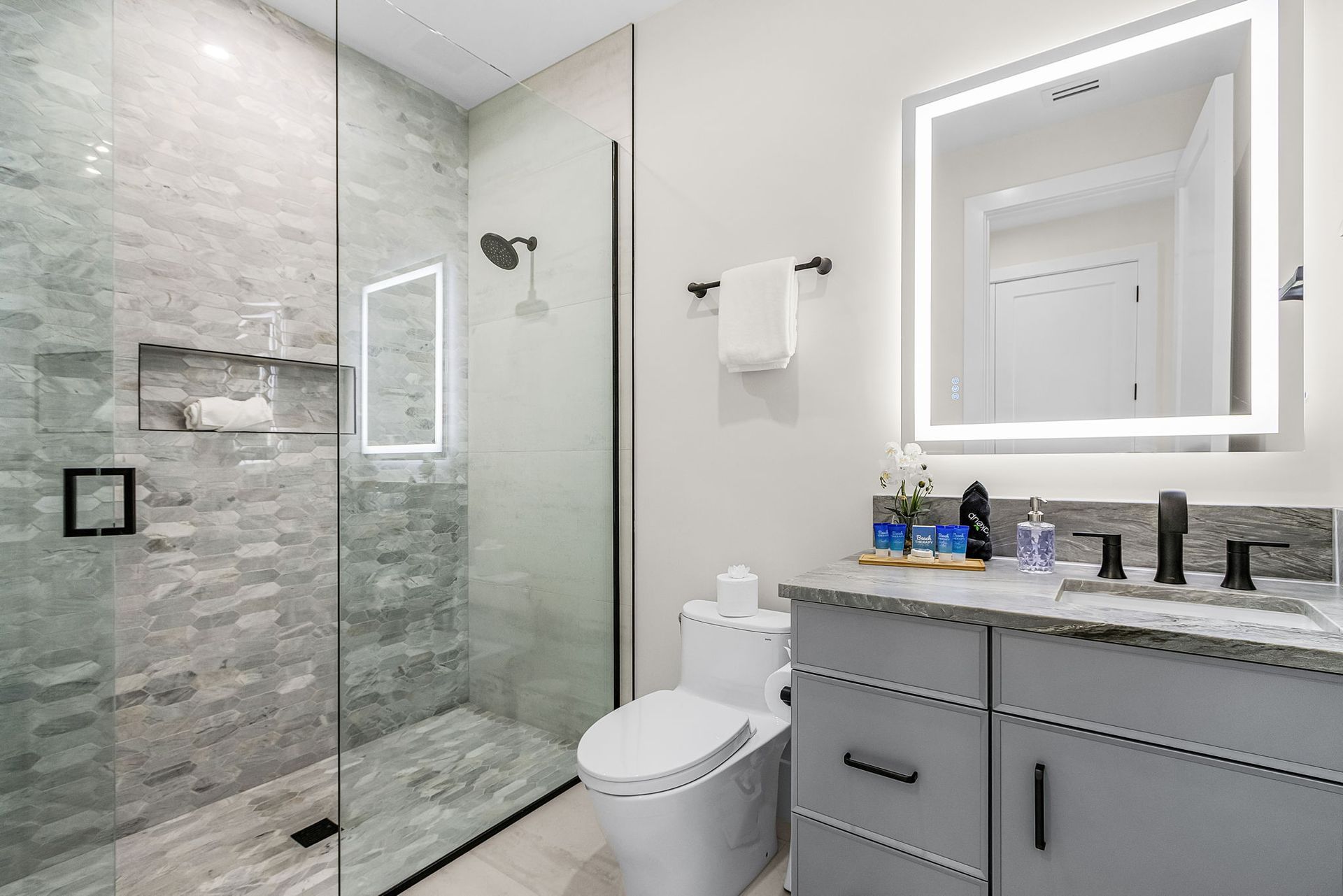 Modern bathroom with glass shower, light gray vanity, toilet, and lit mirror.
