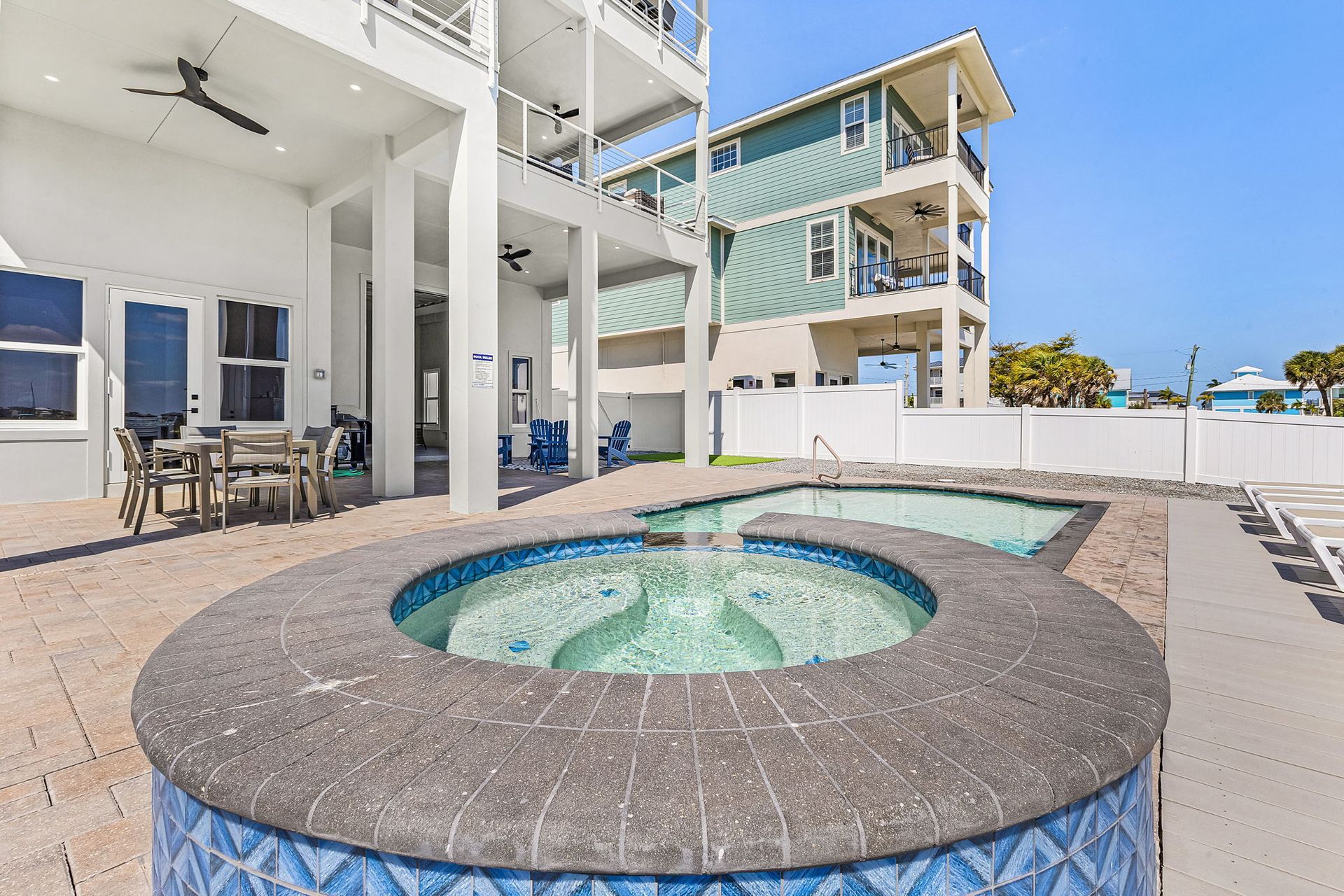Outdoor hot tub beside a white beachside resort, with blue water and patio seating nearby