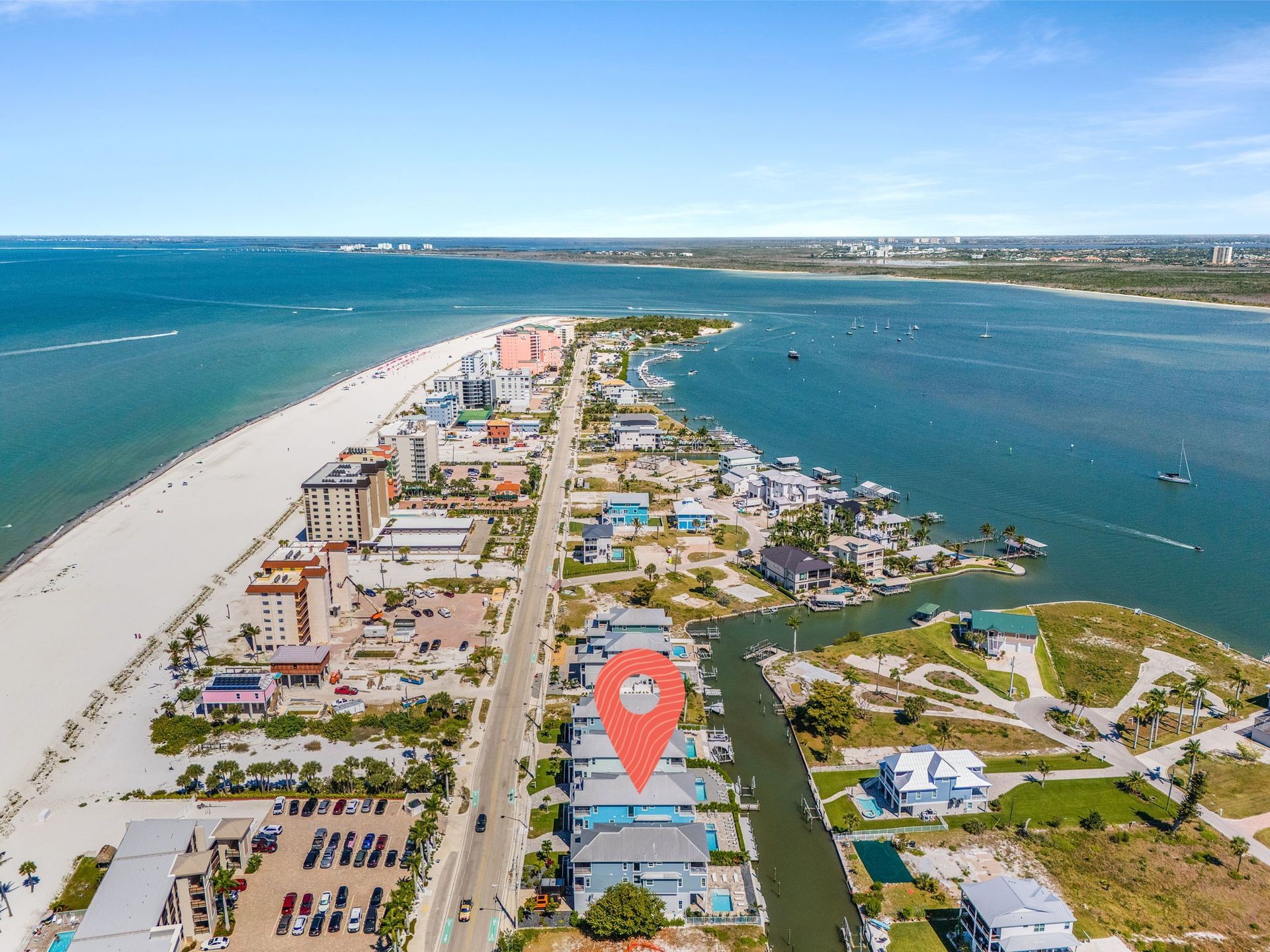 Aerial view of a coastal road between turquoise water and white sand dunes, with homes and a map pin marker