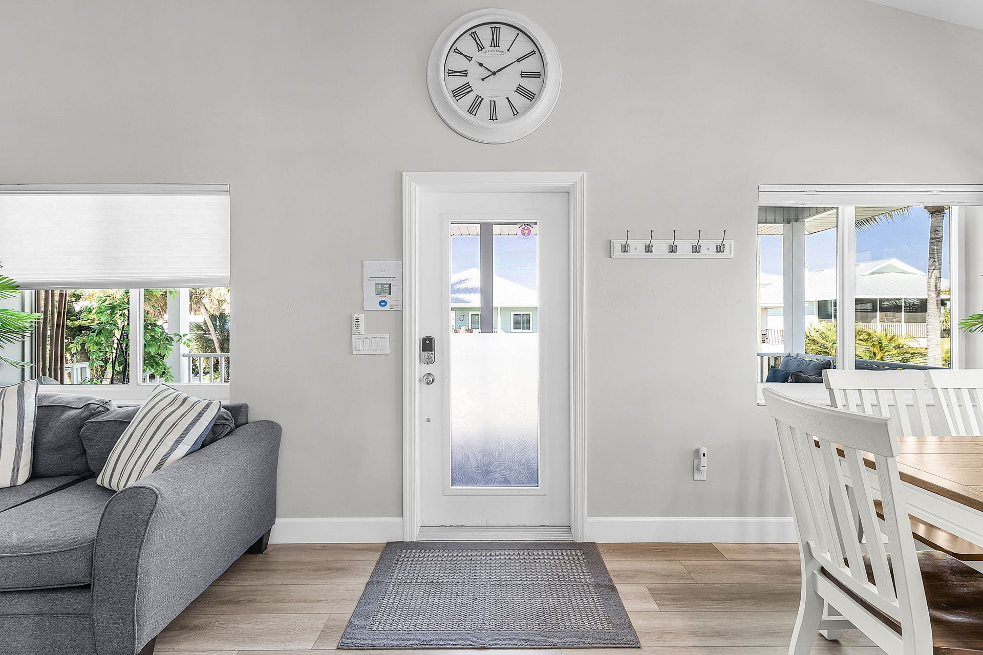 Bright living room with gray sofa, white door and clock, rug, windows, and dining chairs near a patio view