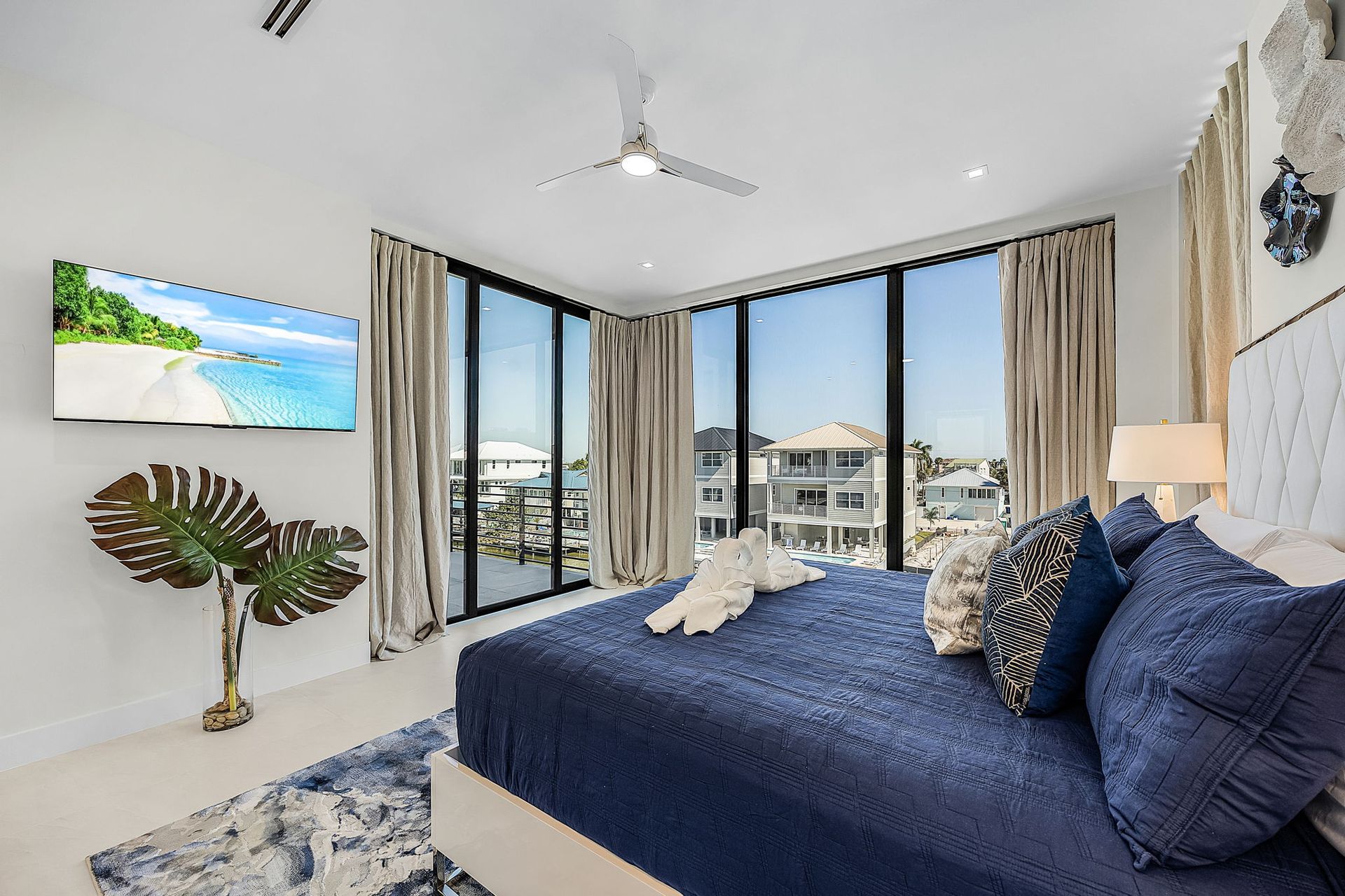 Bedroom with a large bed, TV, tropical plant, and floor-to-ceiling windows overlooking a water view.