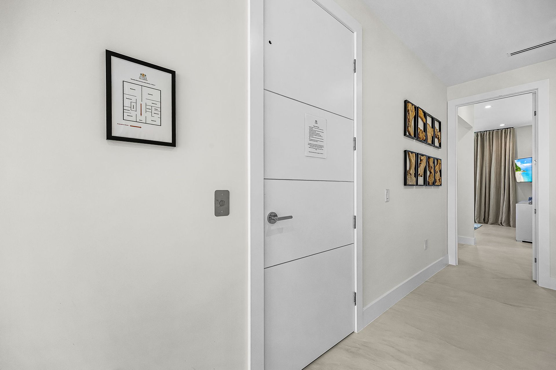 White hallway with closed door, artwork, and an open doorway to a room with curtains and a television.