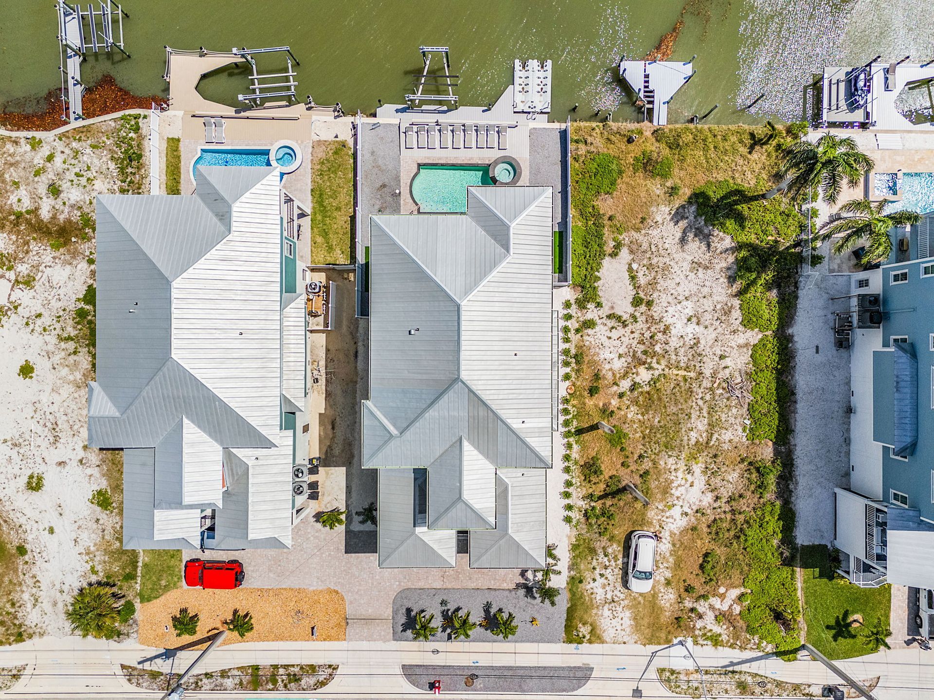 Aerial view of two white-roofed houses with pools, driveways, and adjacent sandy lots