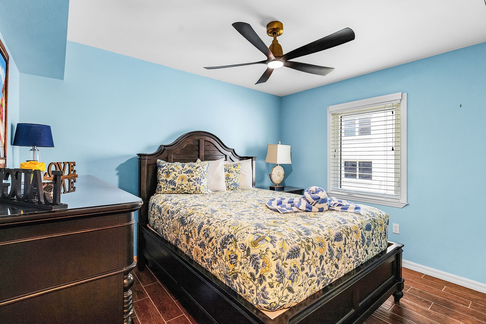Bedroom with dark wood bed, blue walls, floral bedding, dresser, lamp, ceiling fan, and window blinds.