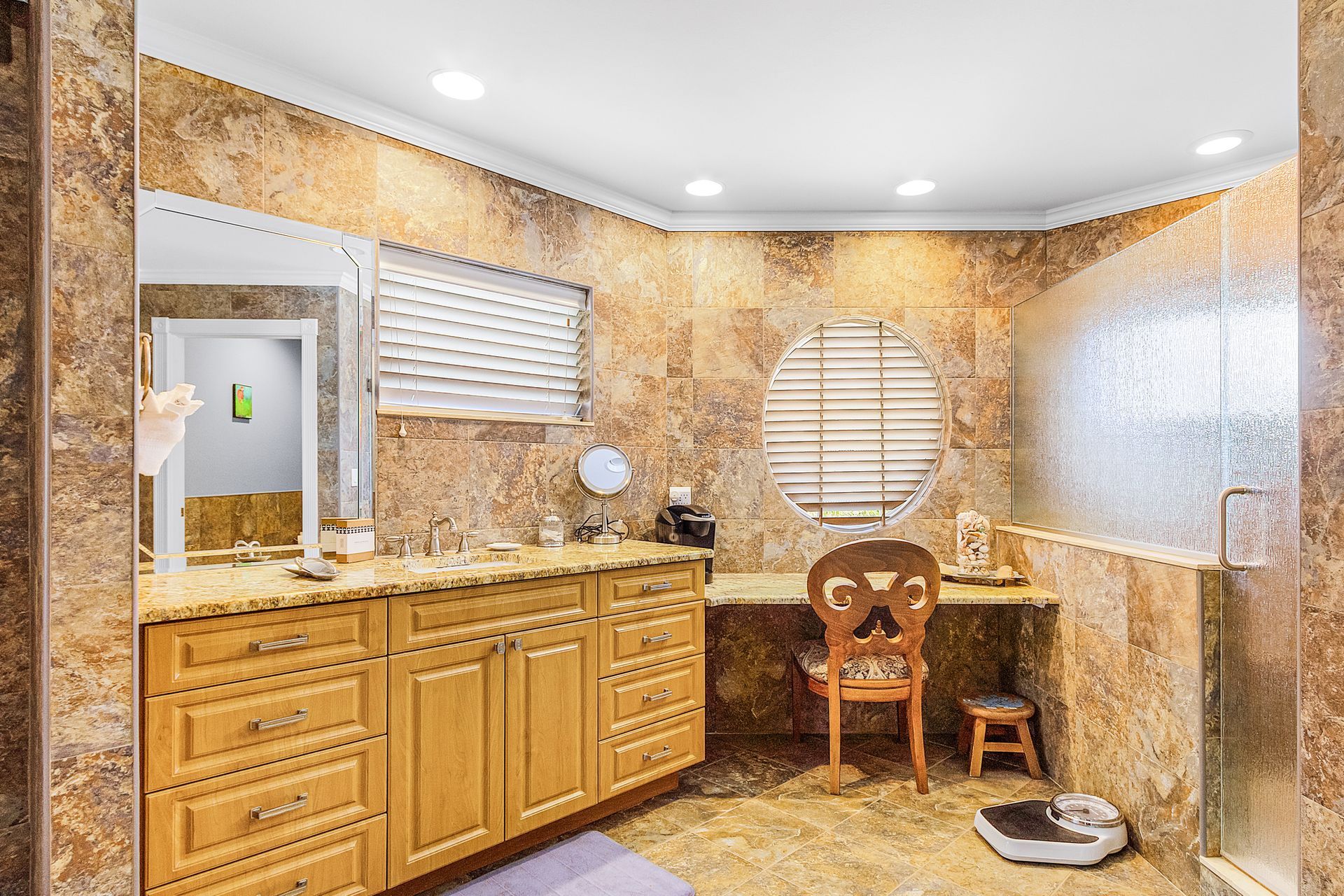 Spacious bathroom with wood cabinets, large mirror, window blinds, vanity, chair, and pet bowl on tiled floor