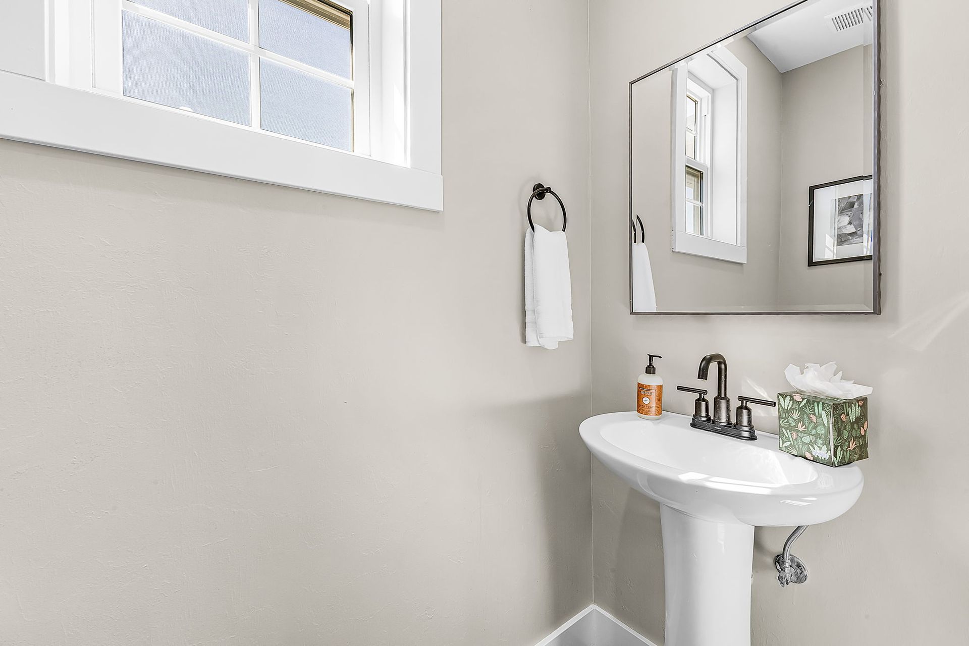 Bright white bathroom with a pedestal sink, mirror, and window on a light gray wall.
