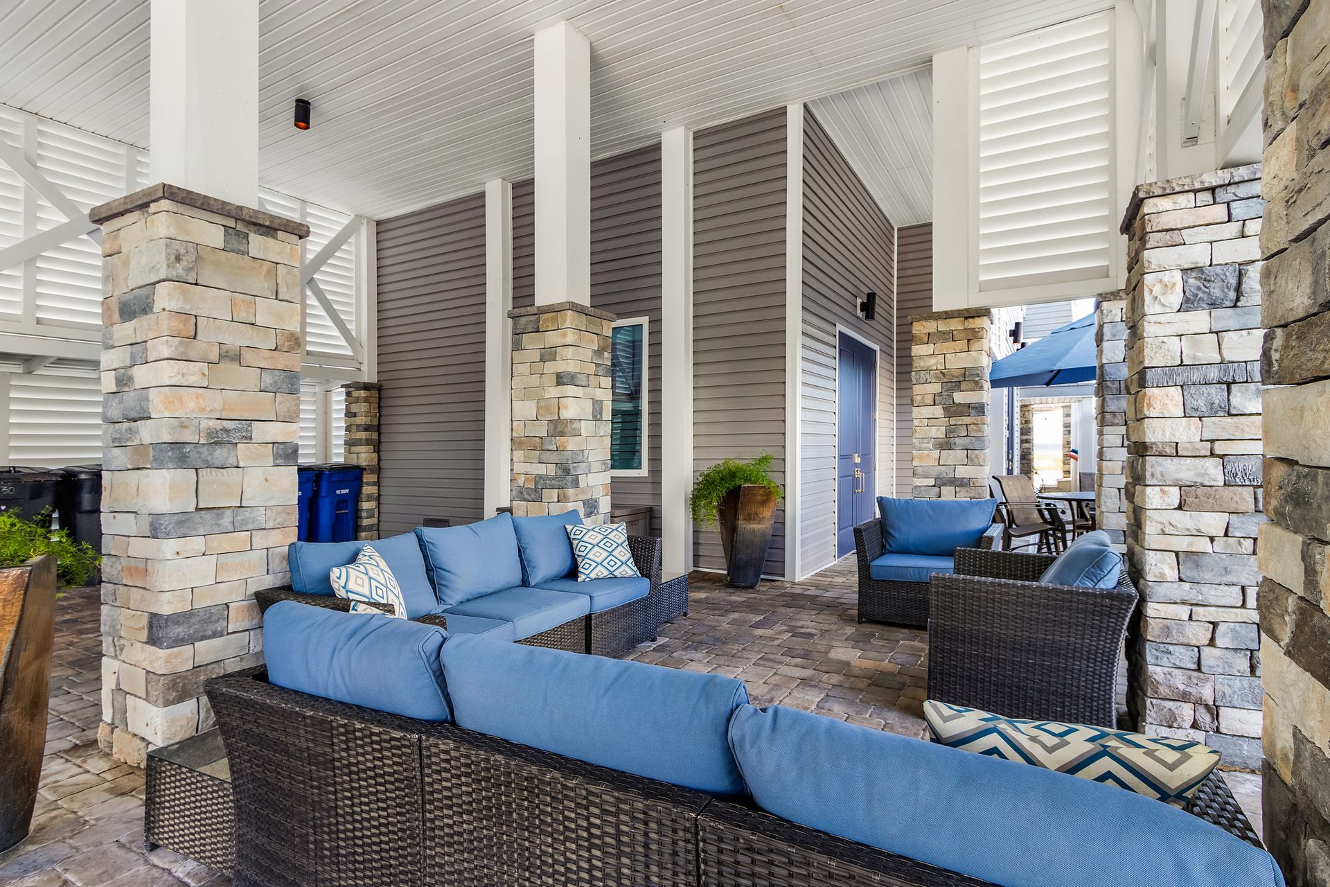 Covered outdoor patio with wicker seating, blue cushions, stone pillars, and a brick fireplace