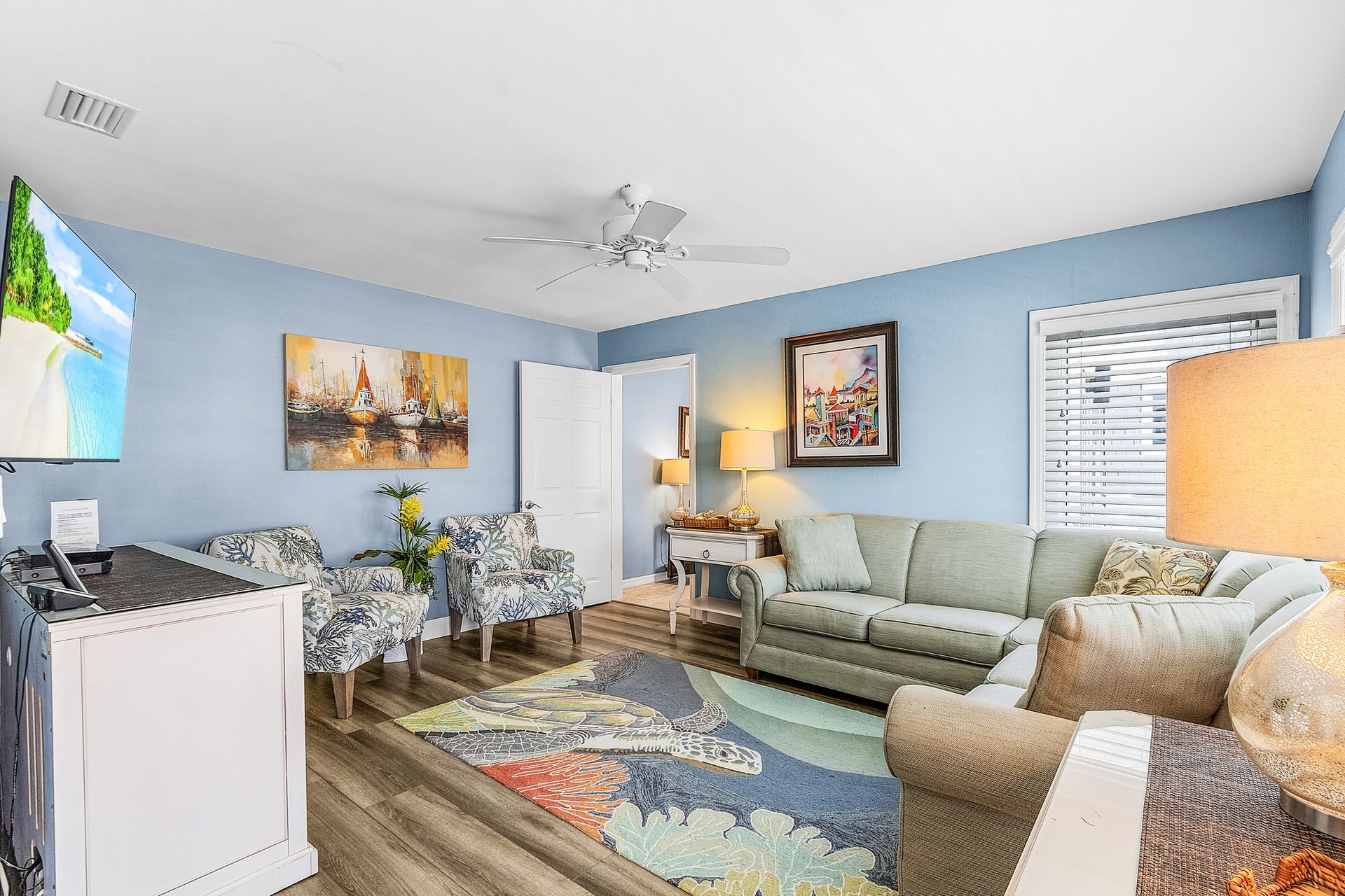 Bright living room with blue walls, green sofa, armchair, rug, lamp, wall art, and a window with blinds