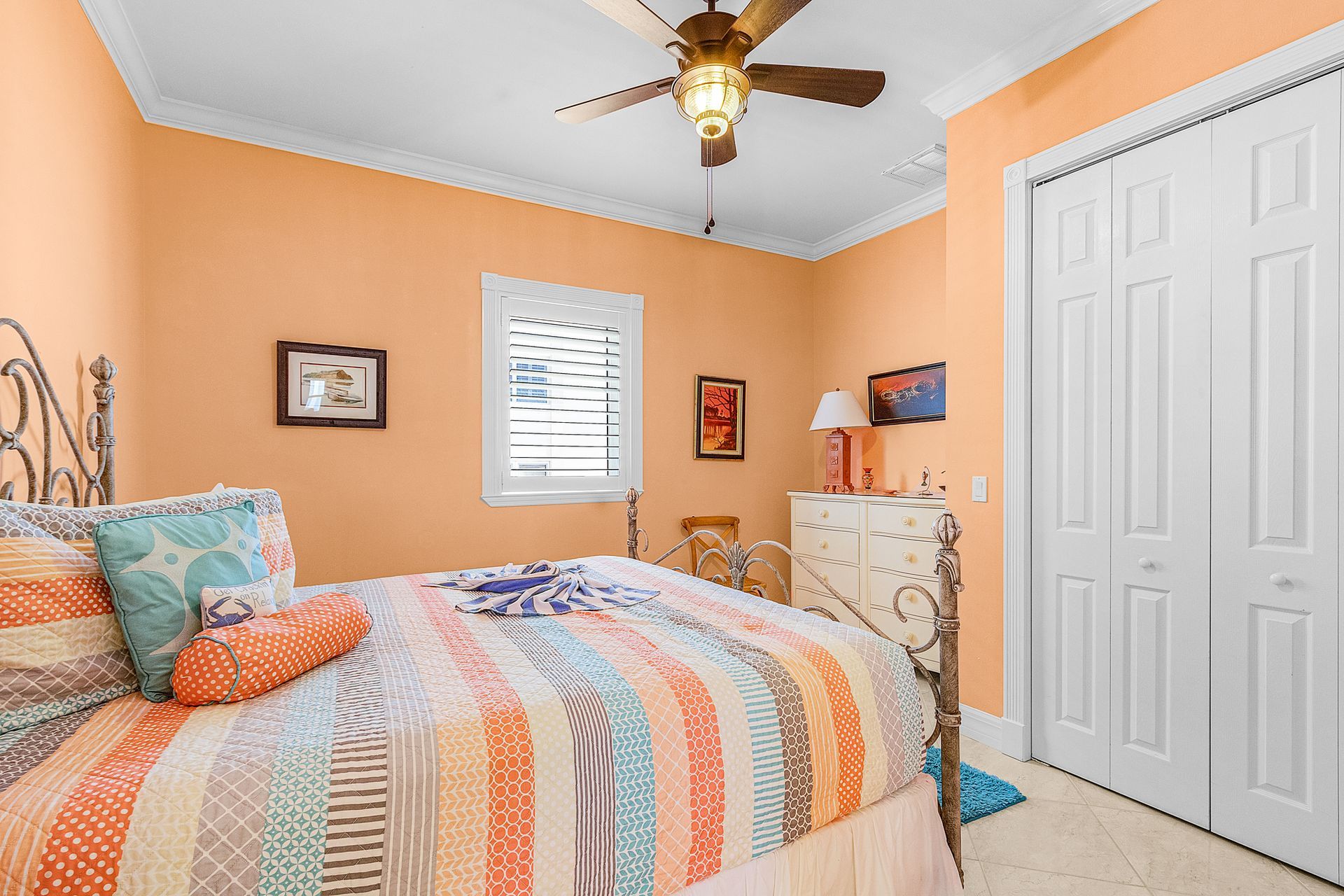 Cozy bedroom with peach walls, colorful striped bedspread, ceiling fan, and white closet doors.