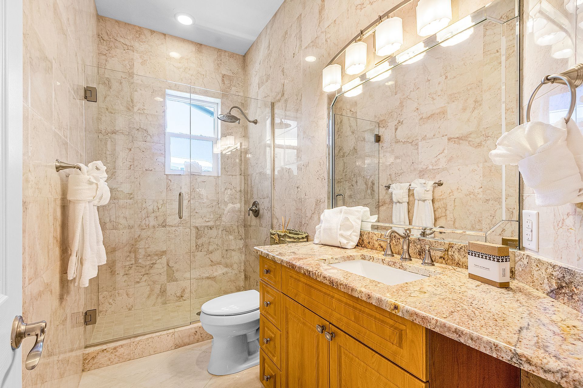 Bright bathroom with beige tile, glass shower, wooden vanity, and toilet.