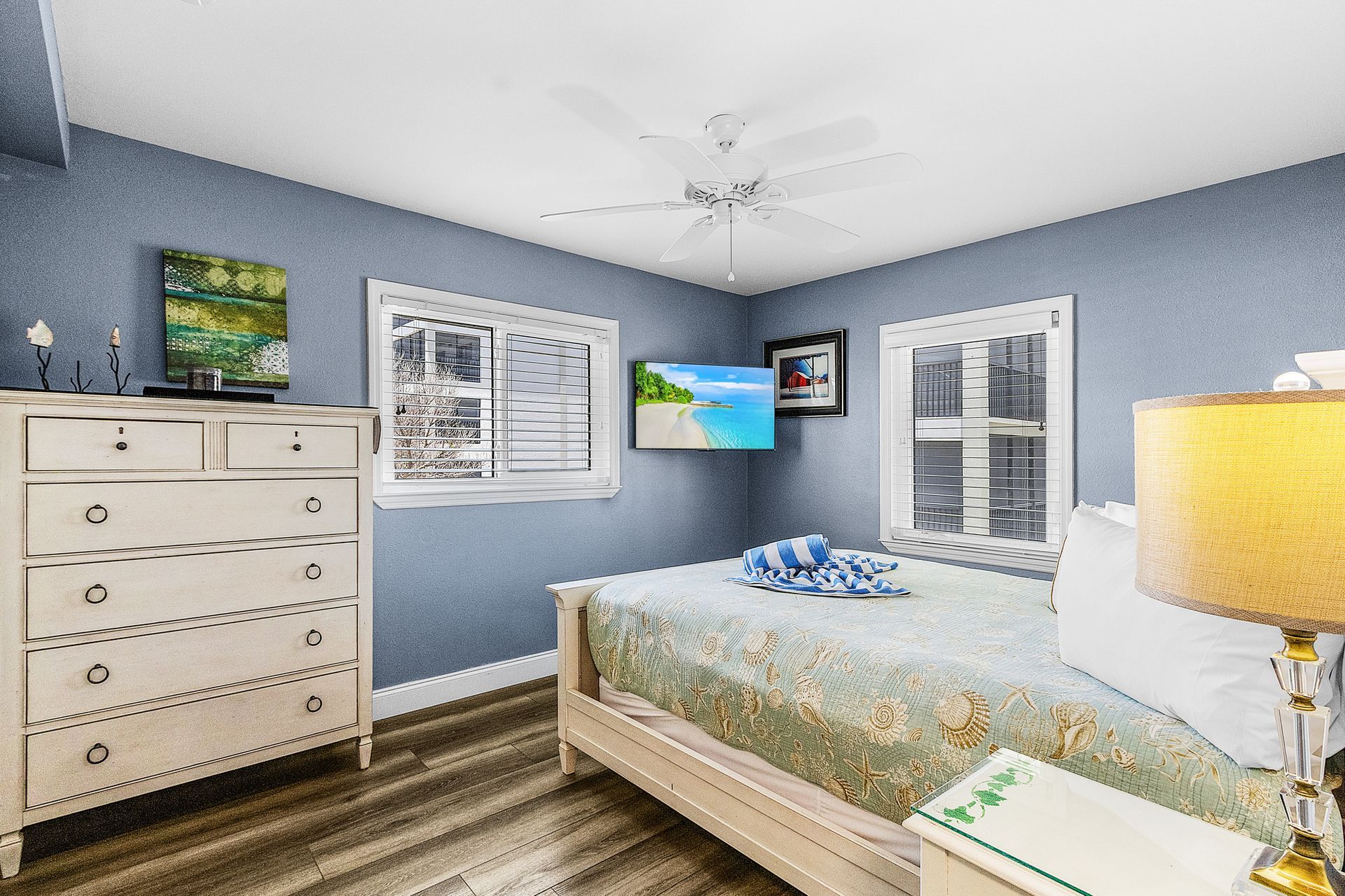 Blue bedroom with a white bed, dresser, lamp, and wall-mounted TV by the window and patio doors