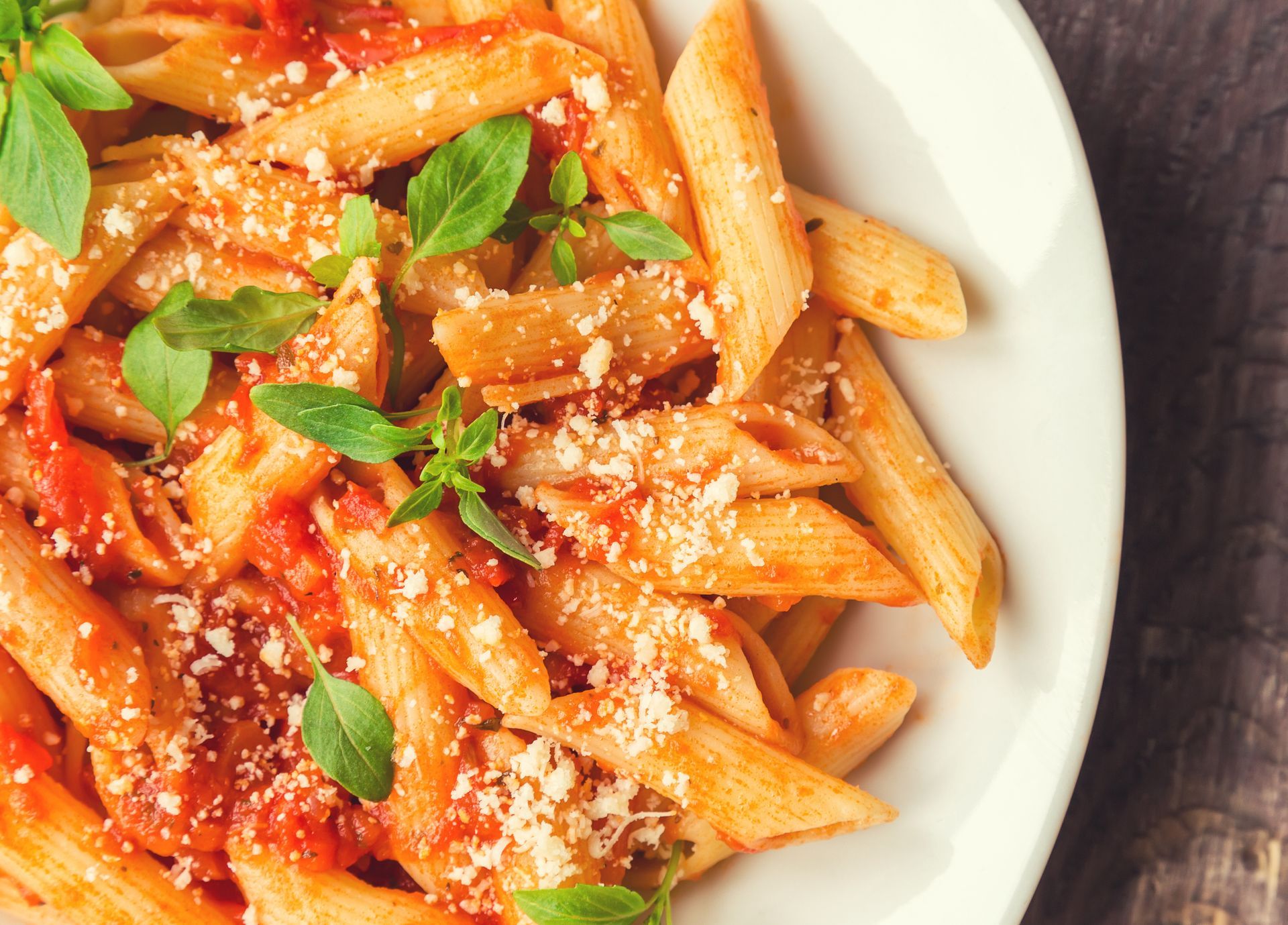 Penne pasta in tomato sauce, garnished with basil and parmesan cheese, in a white bowl.