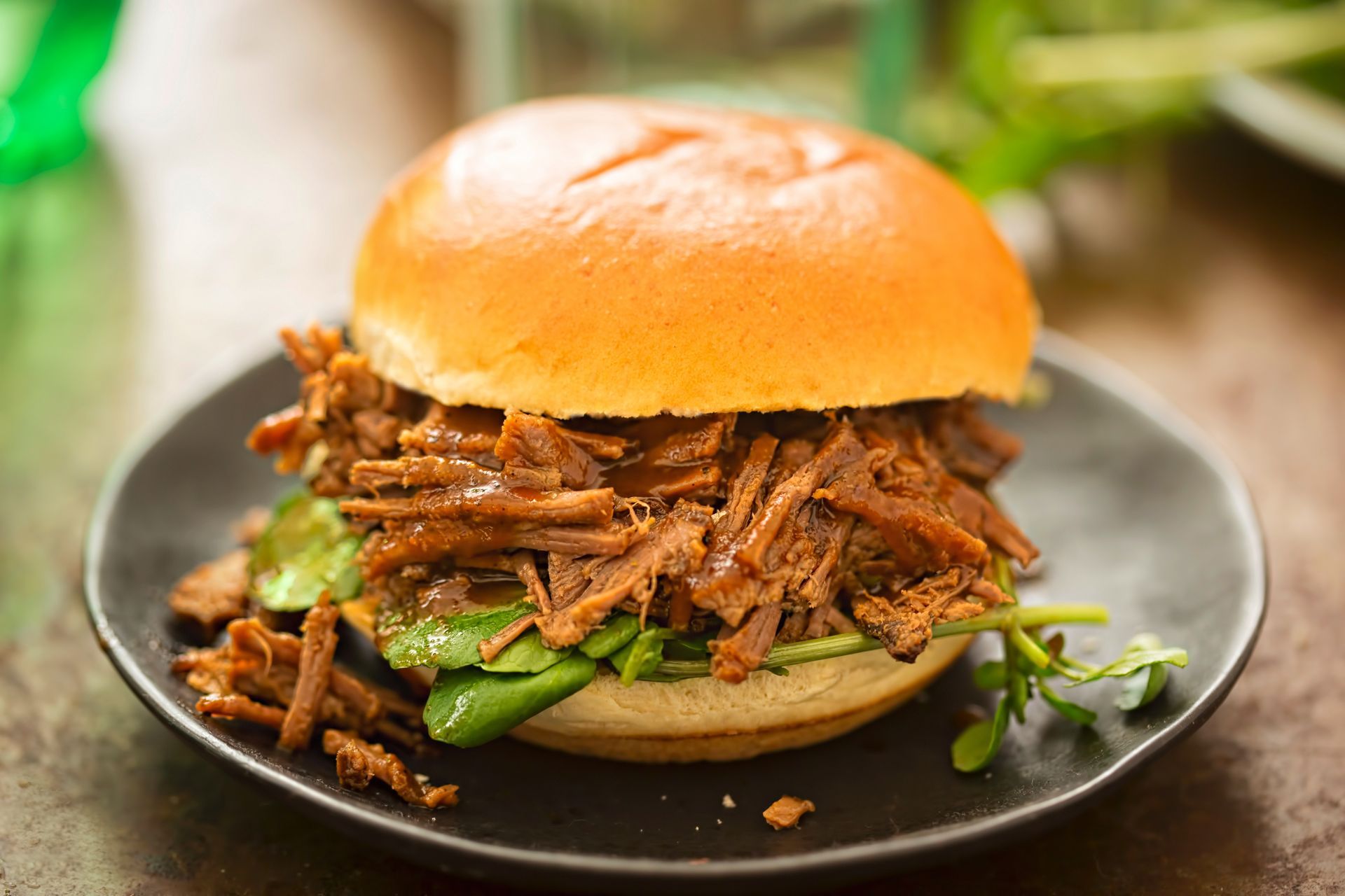 Pulled beef sandwich on a black plate, topped with greens, inside a brioche bun.