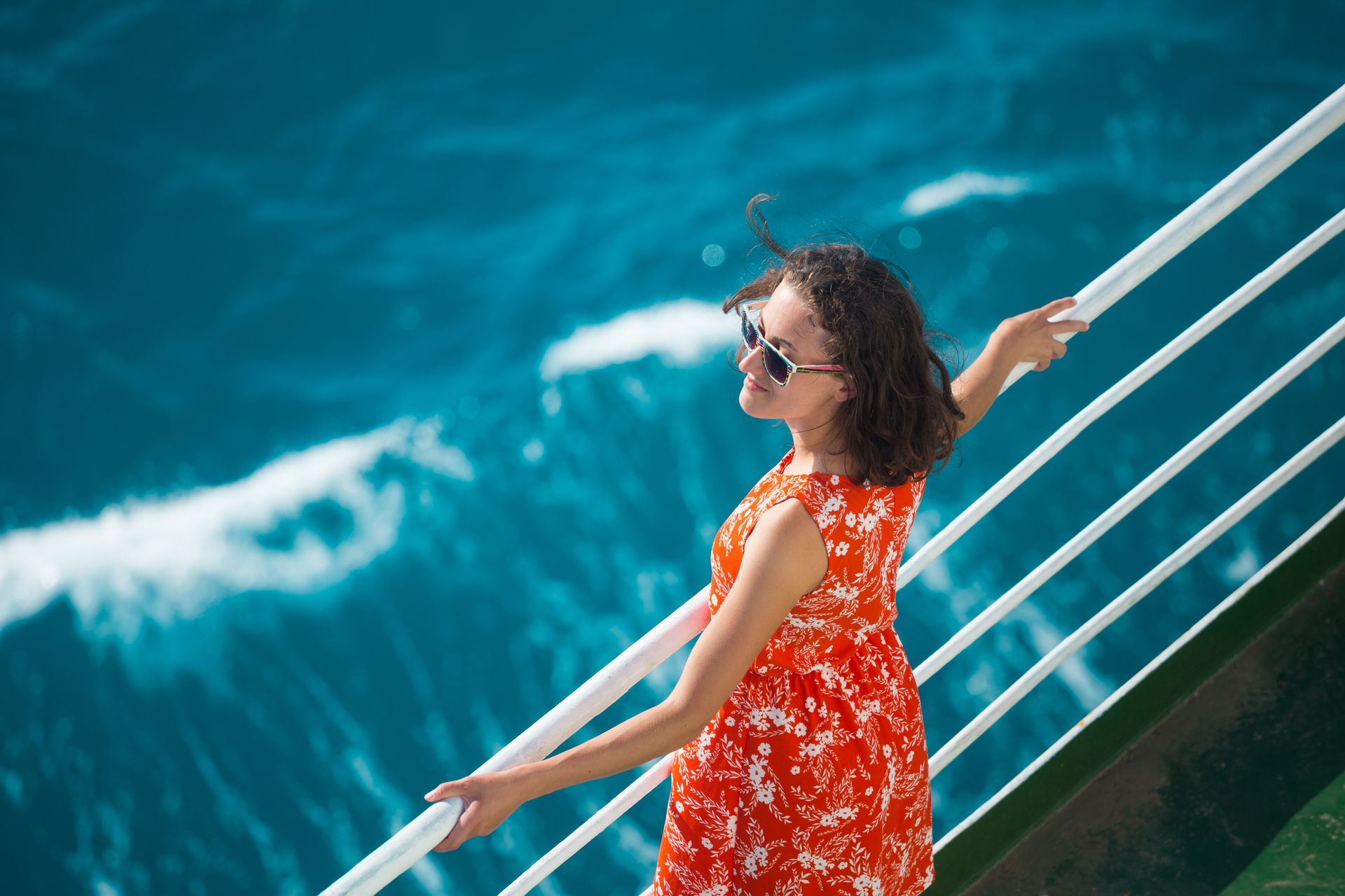 Woman wearing sunglasses and a red dress on a ship, looking at the ocean.