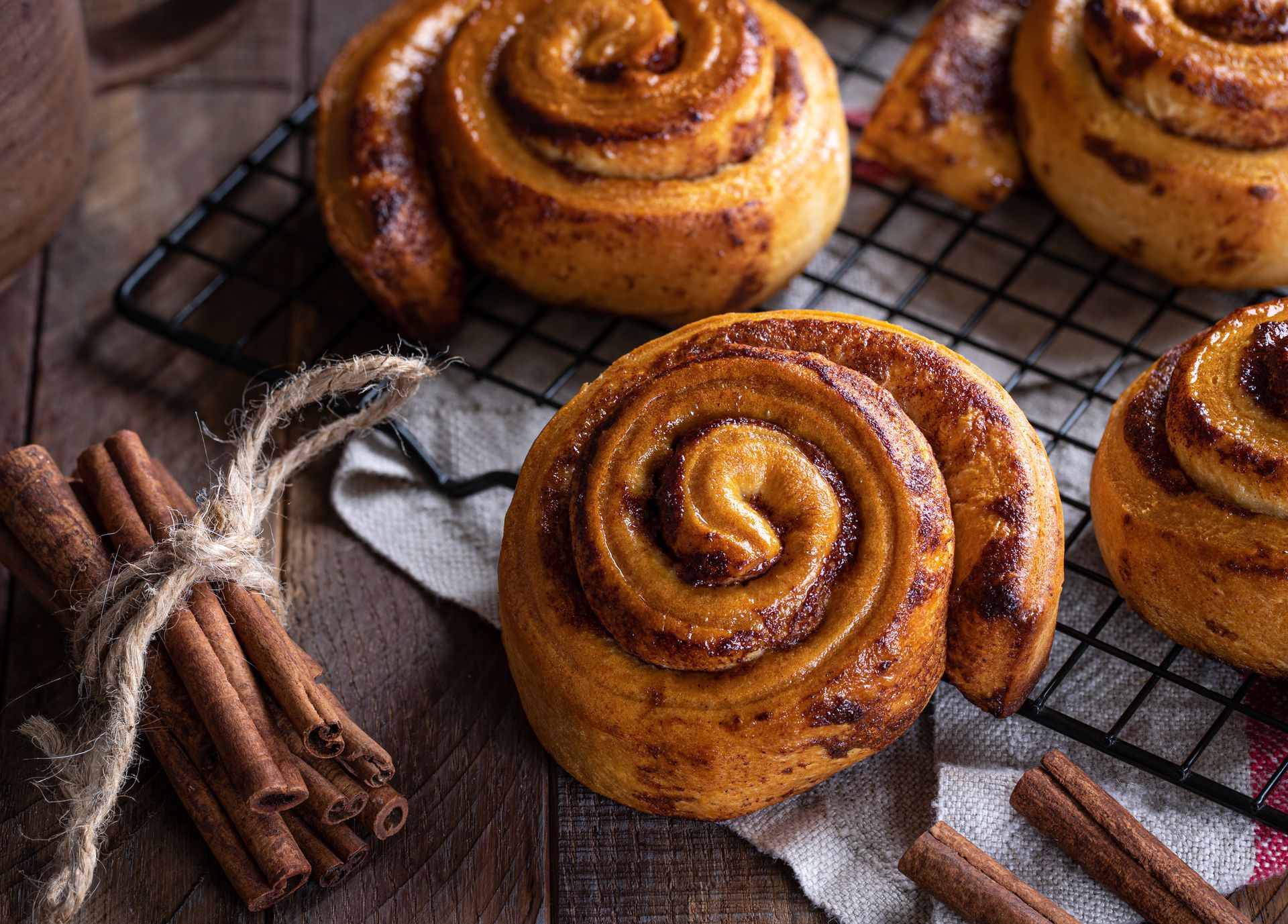Cinnamon rolls on a cooling rack, with cinnamon sticks tied with twine and fabric on a wooden surface.