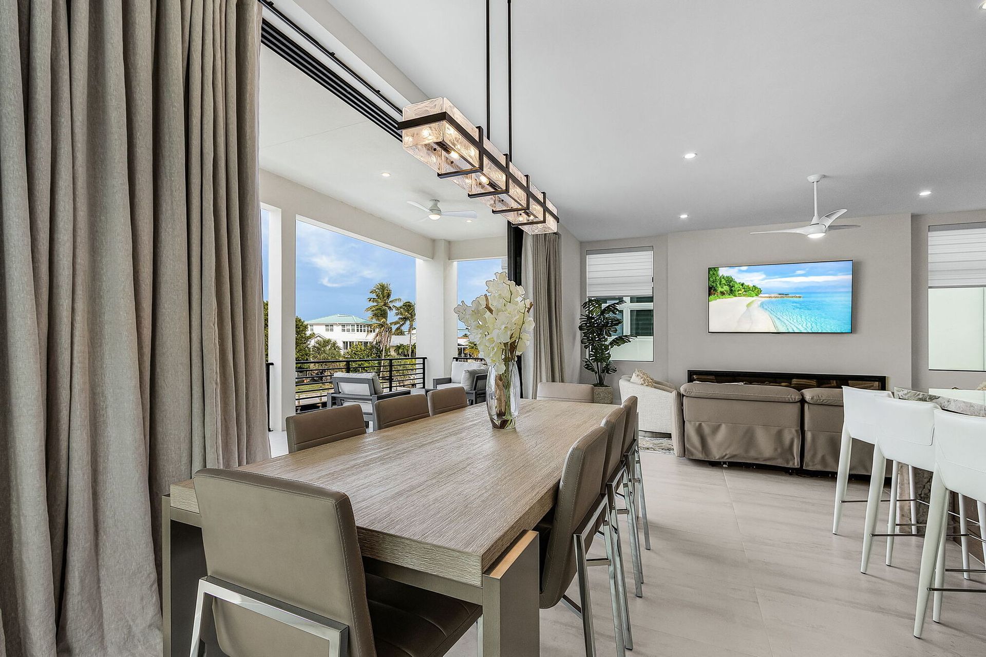 Modern dining area with long table, chairs, large windows, and a wall-mounted TV in a bright open-plan room