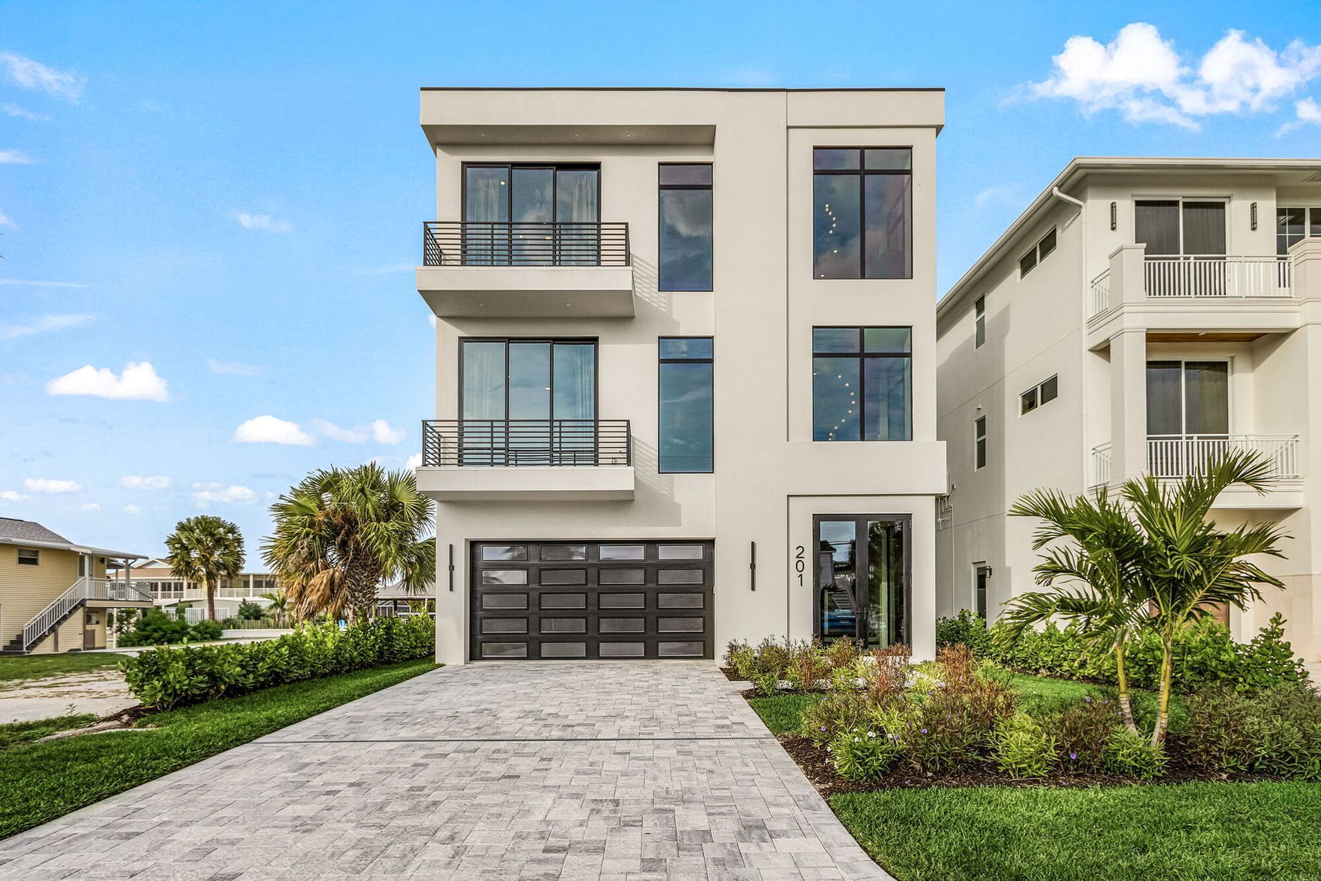 Modern three-story beige house with balcony, garage, and landscaped front yard under a blue sky