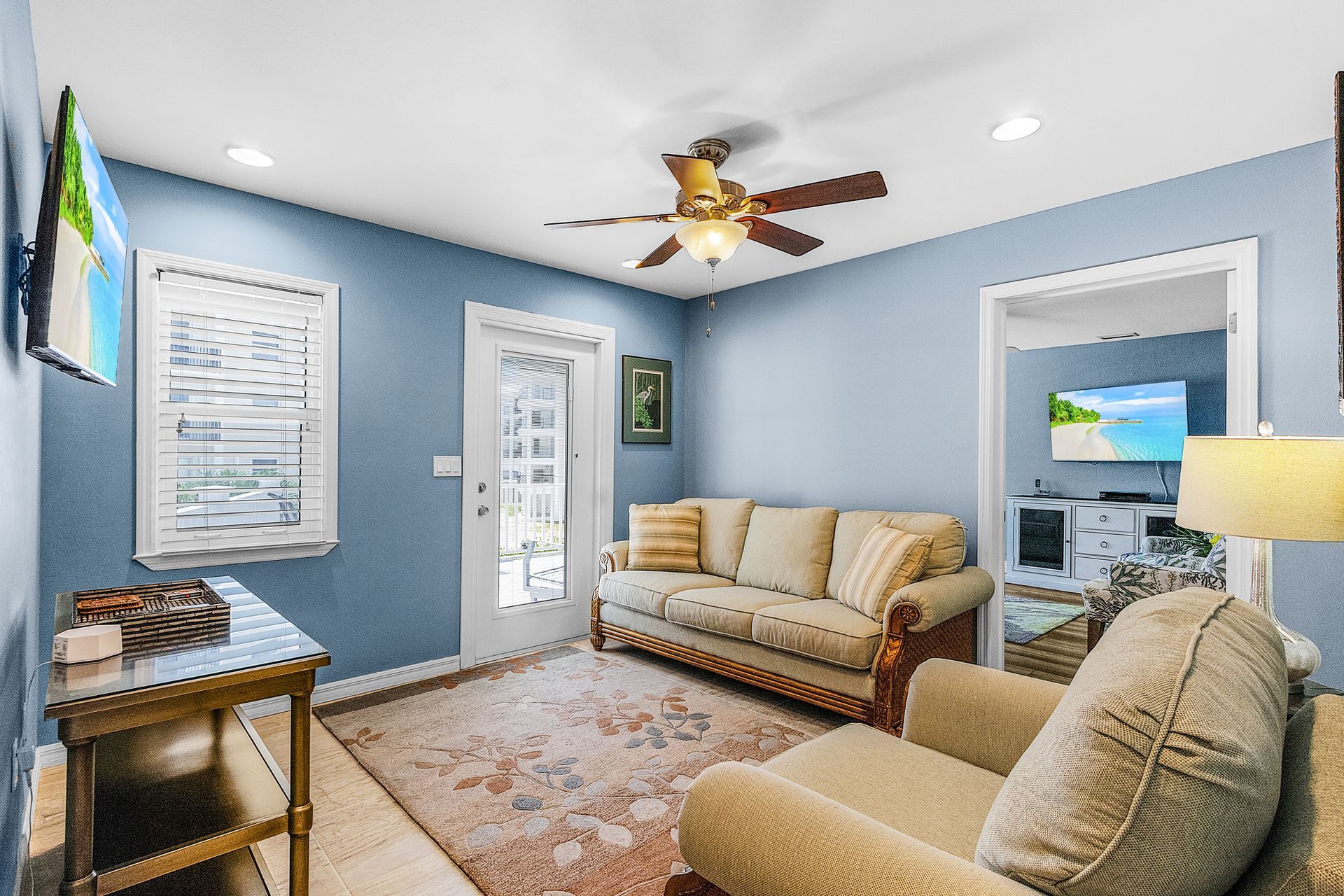 Bright living room with light blue walls, beige sofa and armchair, ceiling fan, and patterned rug.