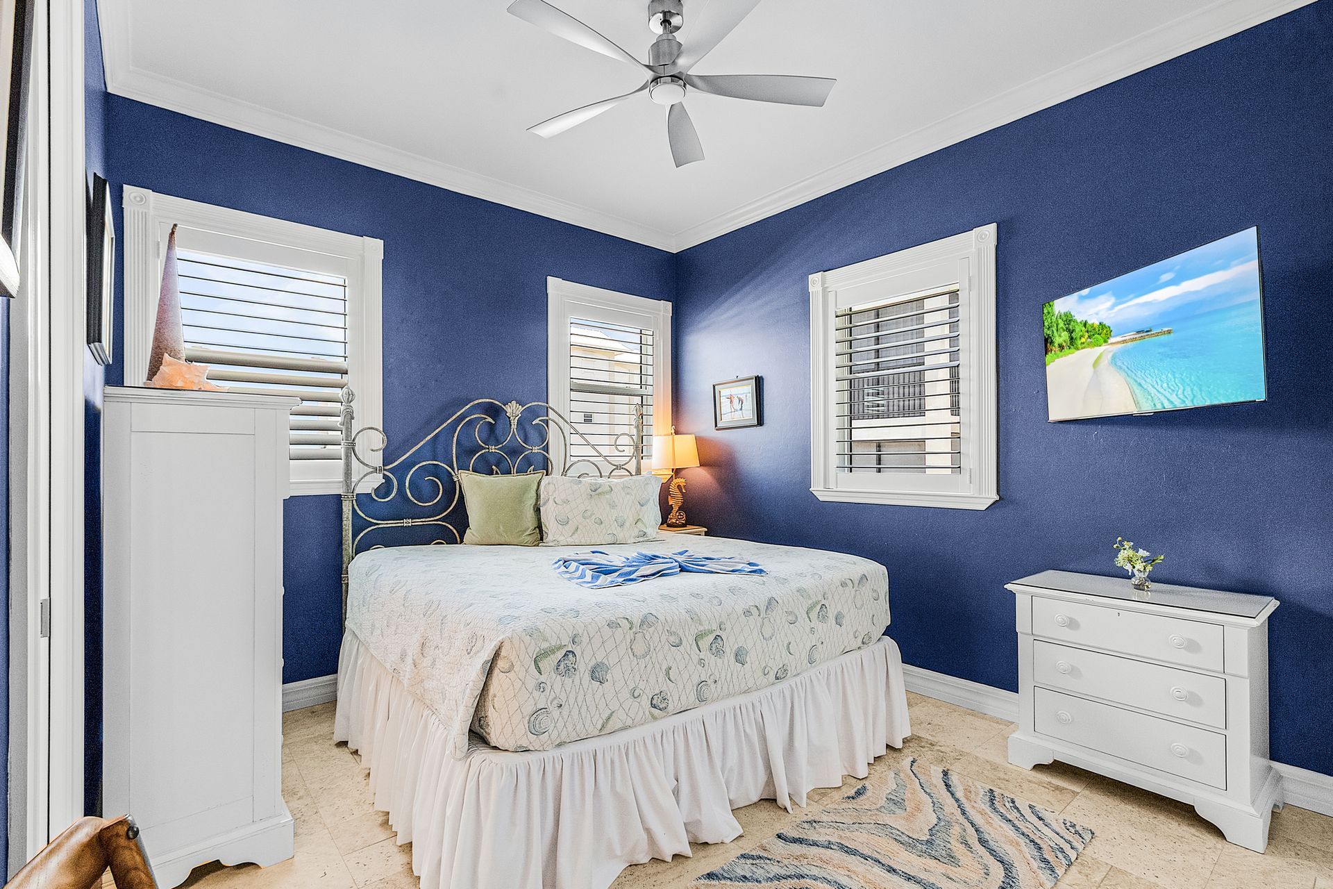 Blue bedroom with wrought-iron bed, white dresser, and bright coastal wall art.