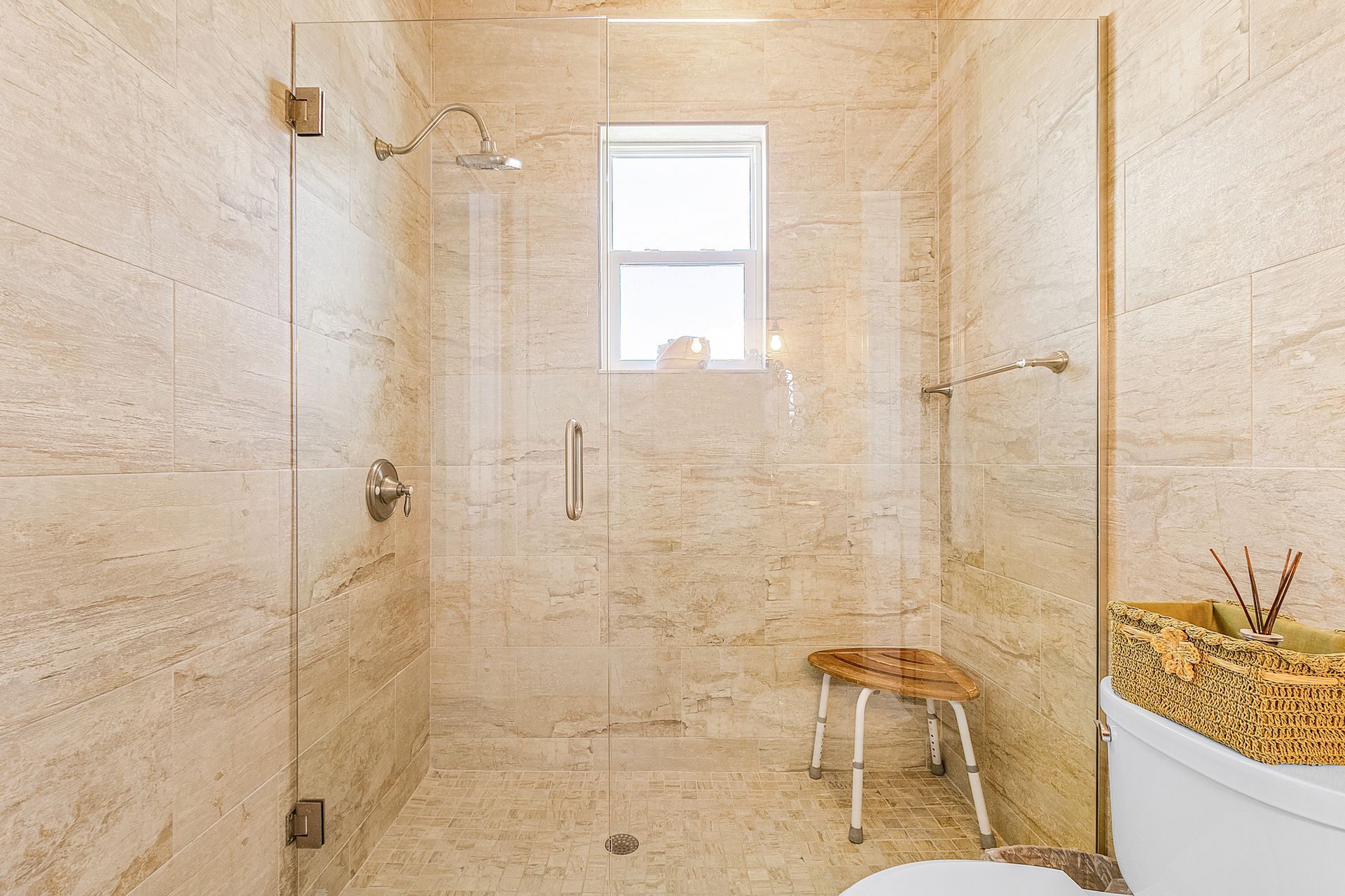 Beige tiled walk-in shower with glass door, bench, window, and woven basket in the bathroom