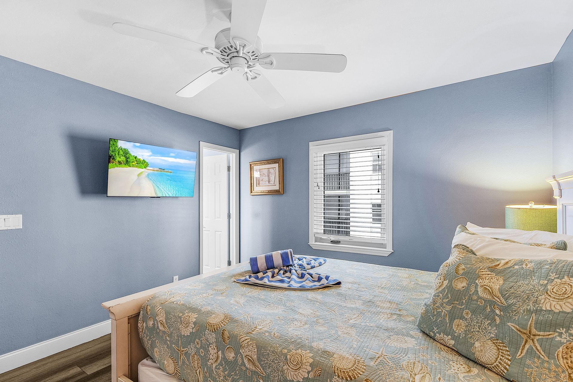 Blue bedroom with bed, wall-mounted TV, window blinds, and ceiling fan; a tray sits on the bed.