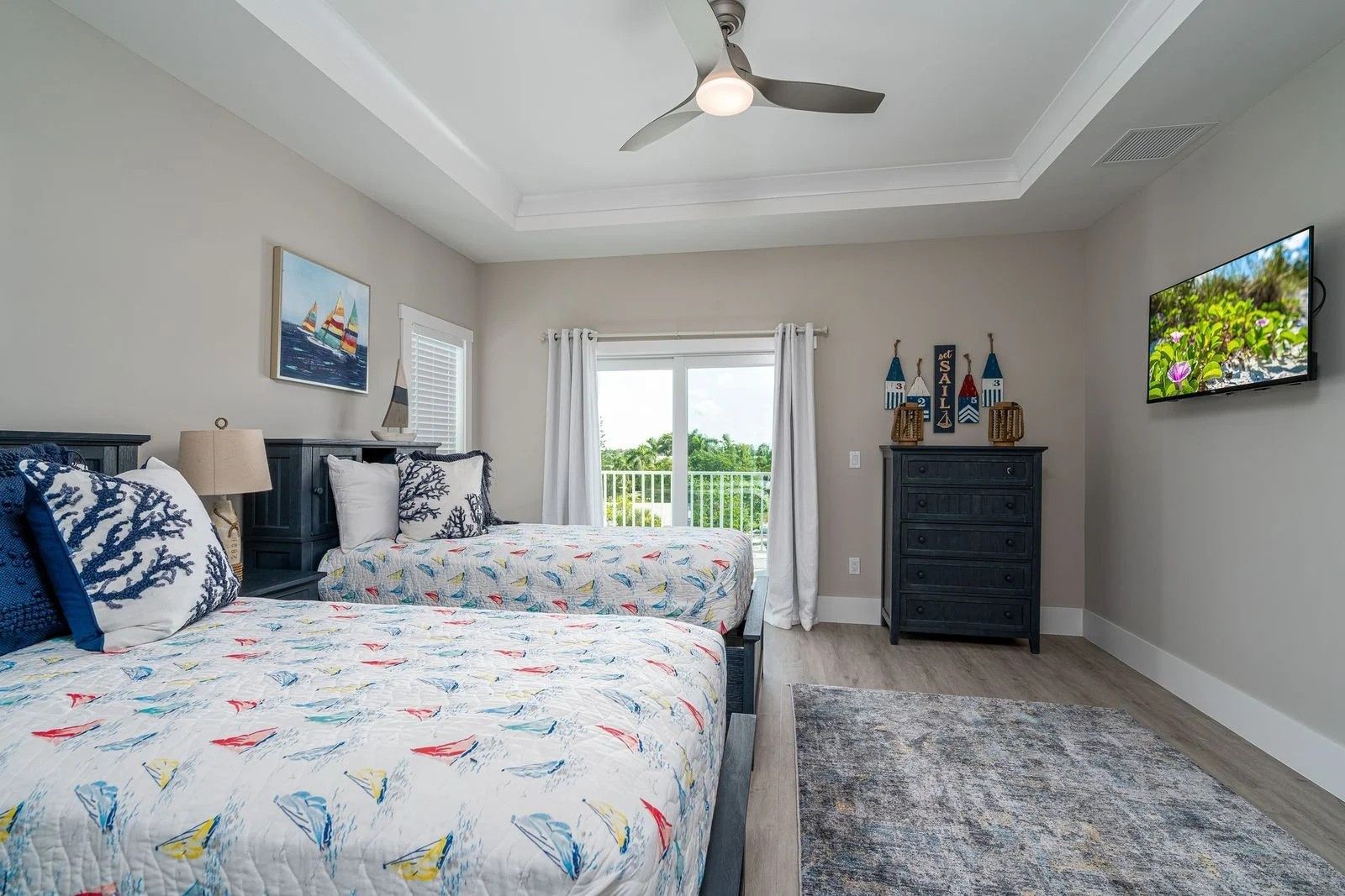 Bright bedroom with bed, ceiling fan, dresser, wall TV, and balcony doors.